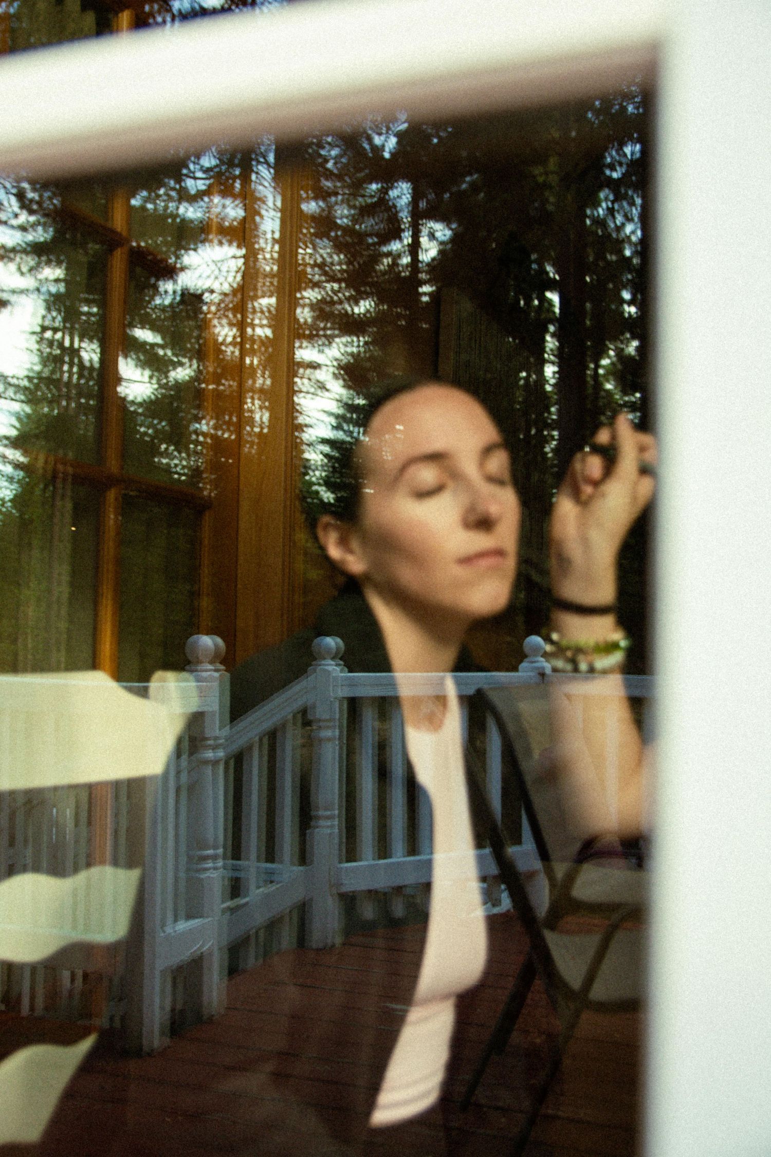 Woman with closed eyes photographed through a window with forest reflections in soft natural light.