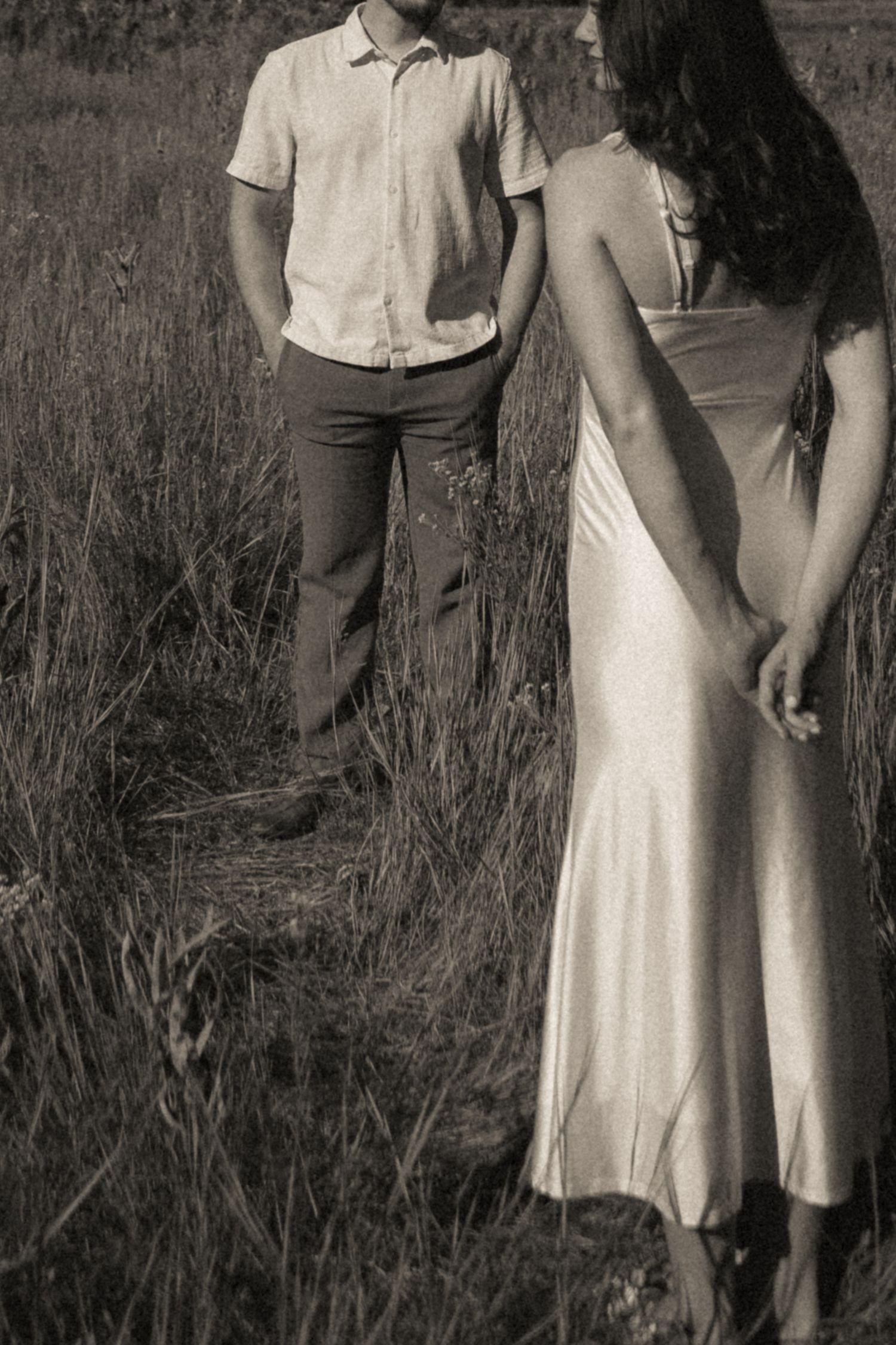 Black and white photo of a couple standing in a grassy field, woman in silk dress facing a man in casual shirt.