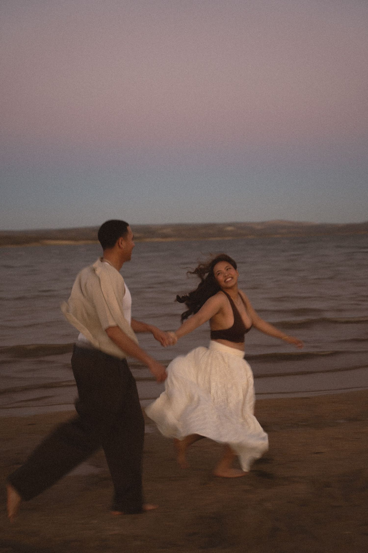 Joyful couple running and holding hands on beach at dusk with ocean behind.