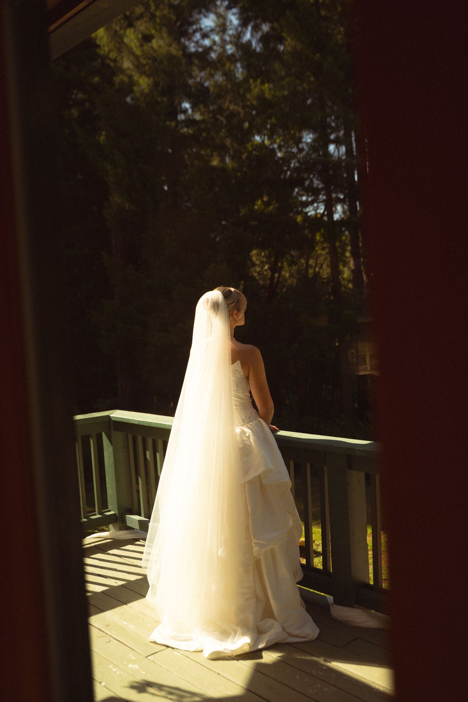 A bride in a flowing white gown and veil stands on a sunlit wooden deck surrounded by tall trees.