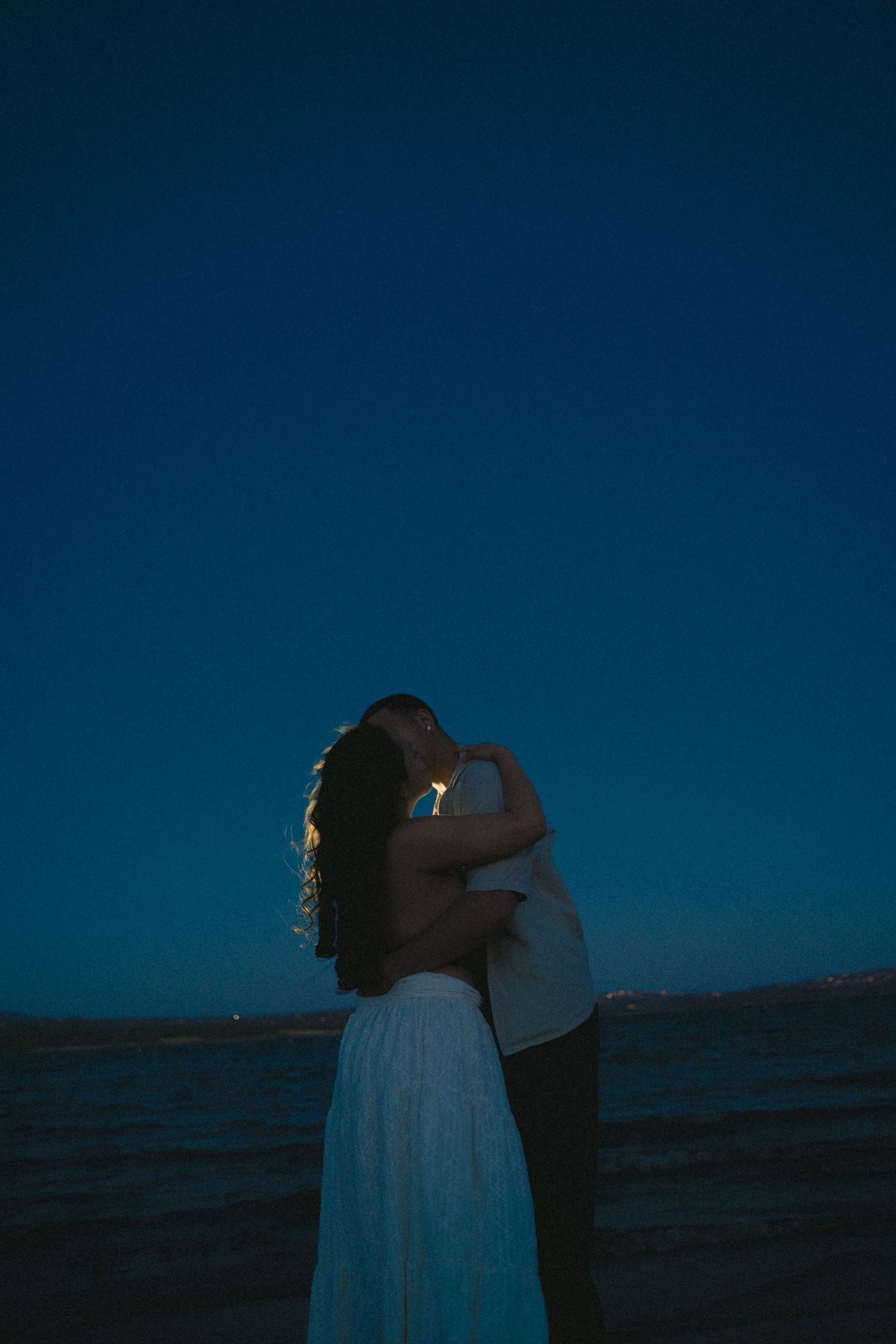 Couple embracing at twilight on a beach under a deep blue evening sky near calm water.