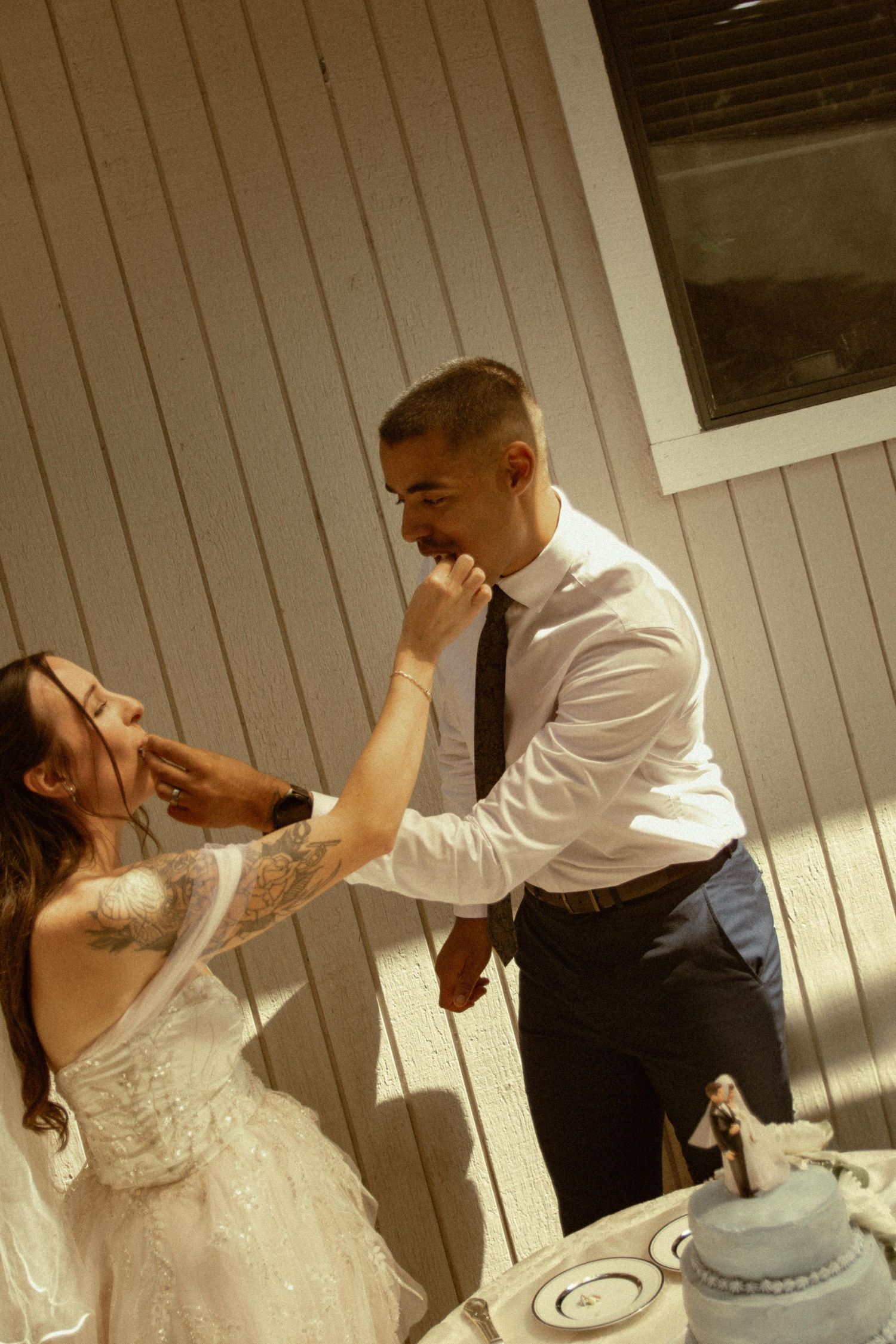 Bride and groom playfully feeding each other wedding cake outdoors in bright sunlight.