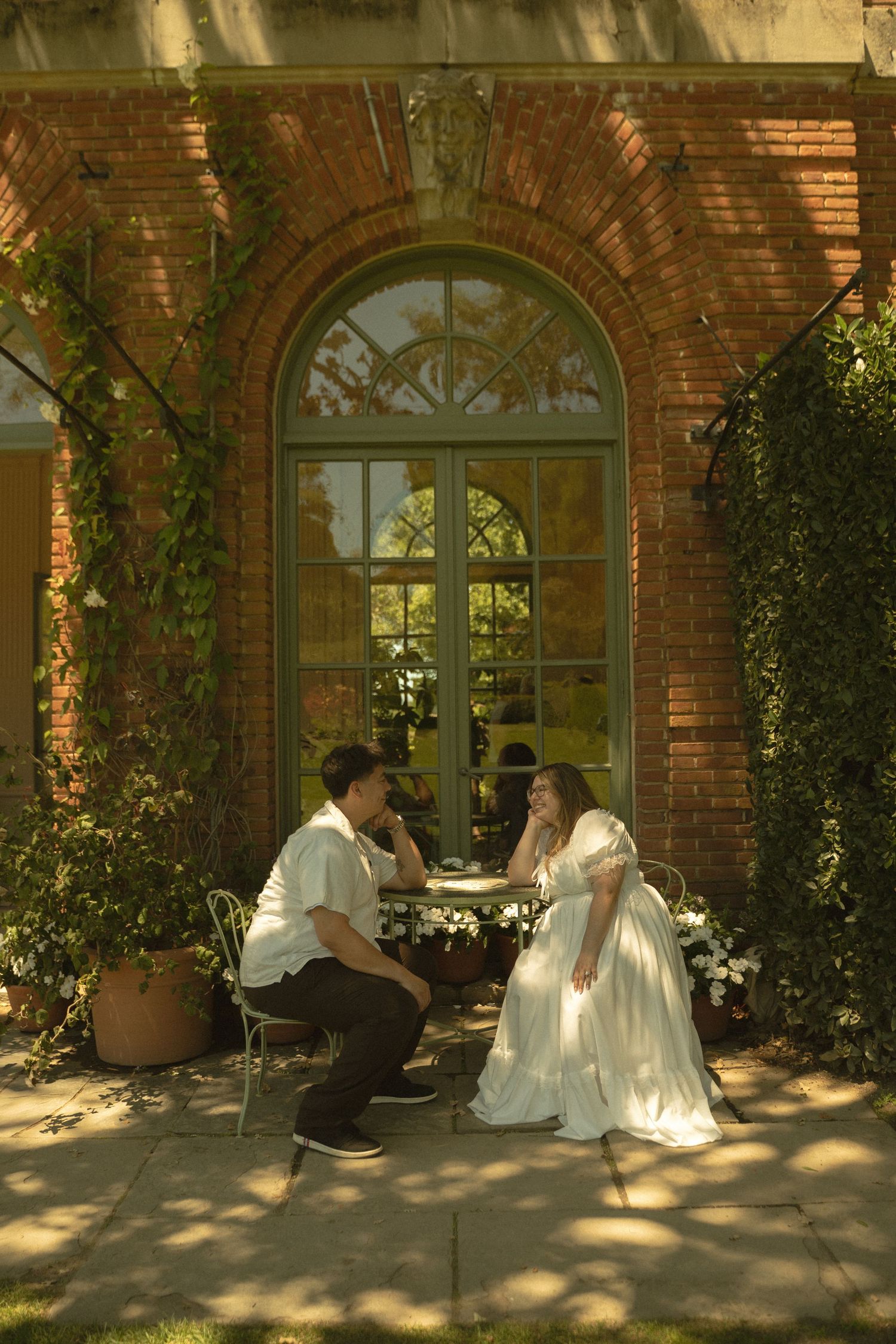 Bride and groom sharing intimate moment at garden table outside elegant brick building with arched window.