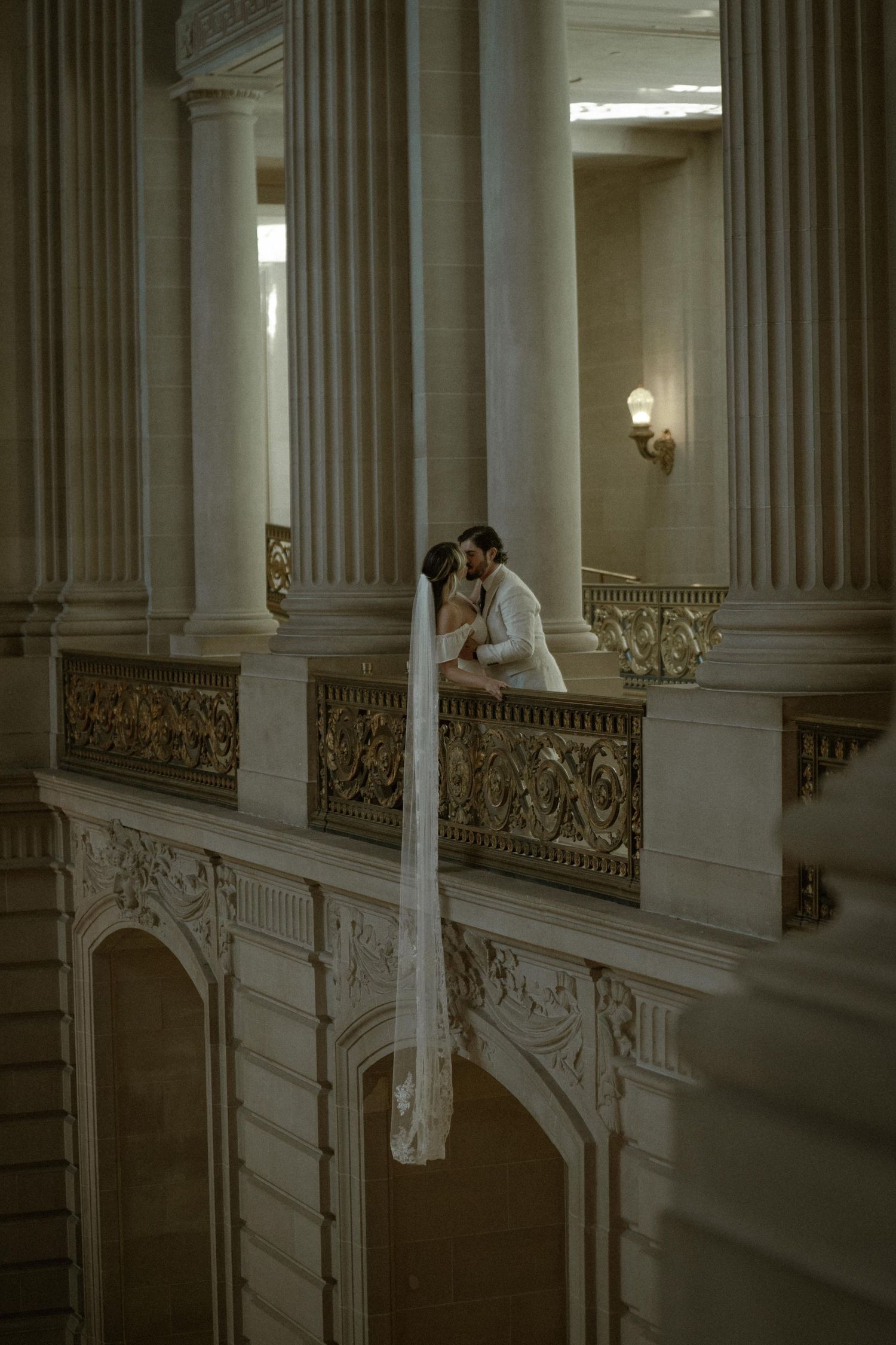Bride and groom share a kiss on an ornate balcony with grand columns at San Francisco City Hall.