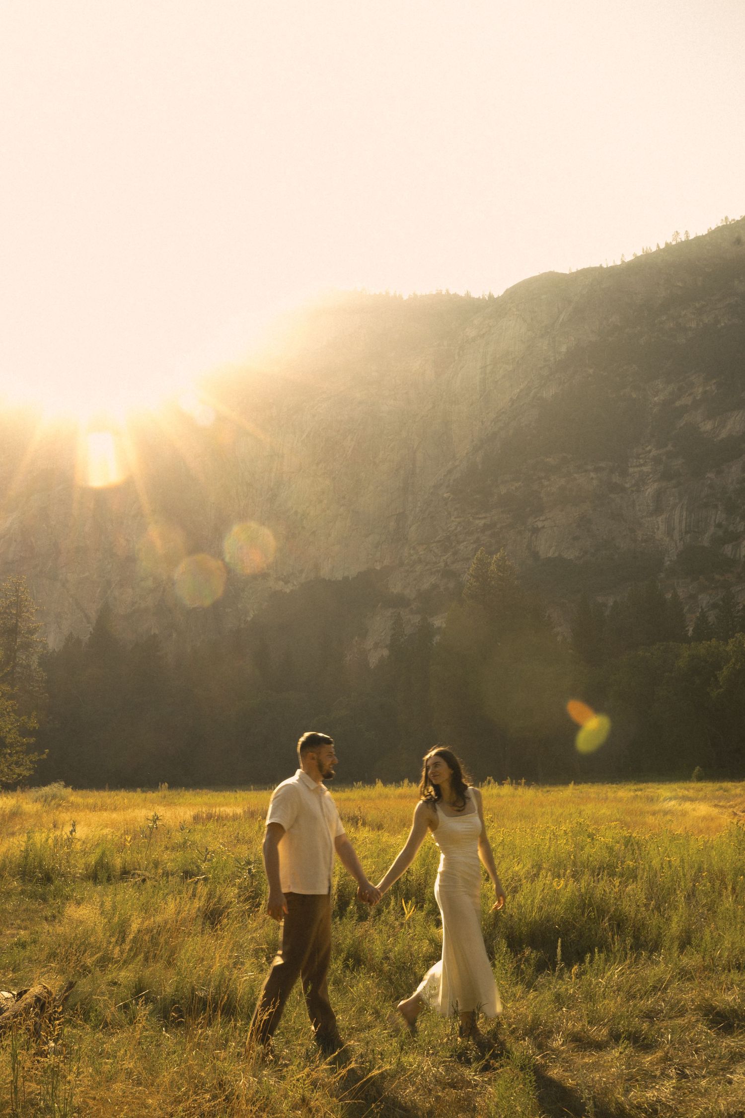 Couple holds hands walking through golden meadow at sunset with mountain backdrop in Yosemite Valley.