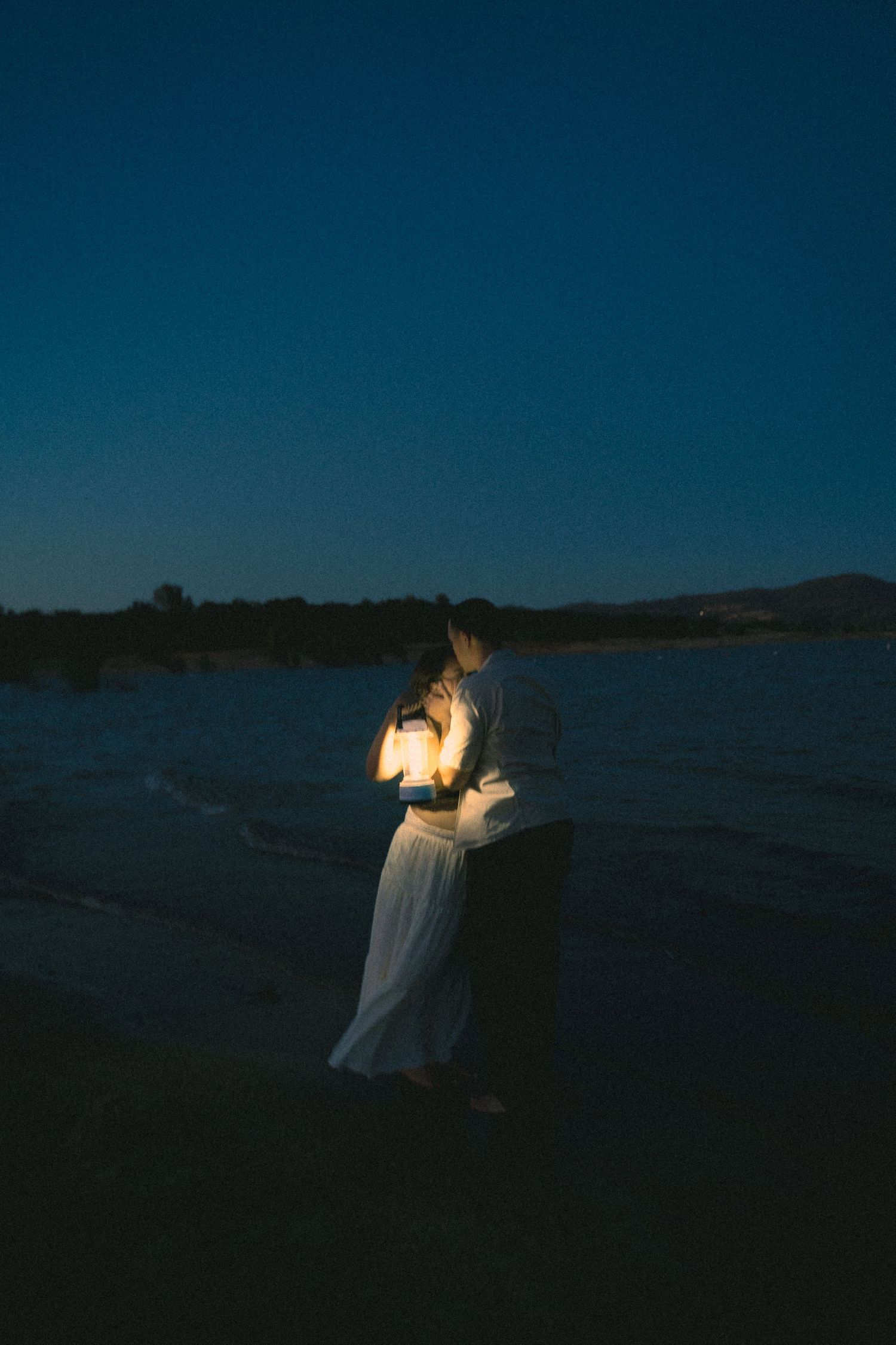 Couple embracing while holding glowing lantern on dark beach under twilight sky.