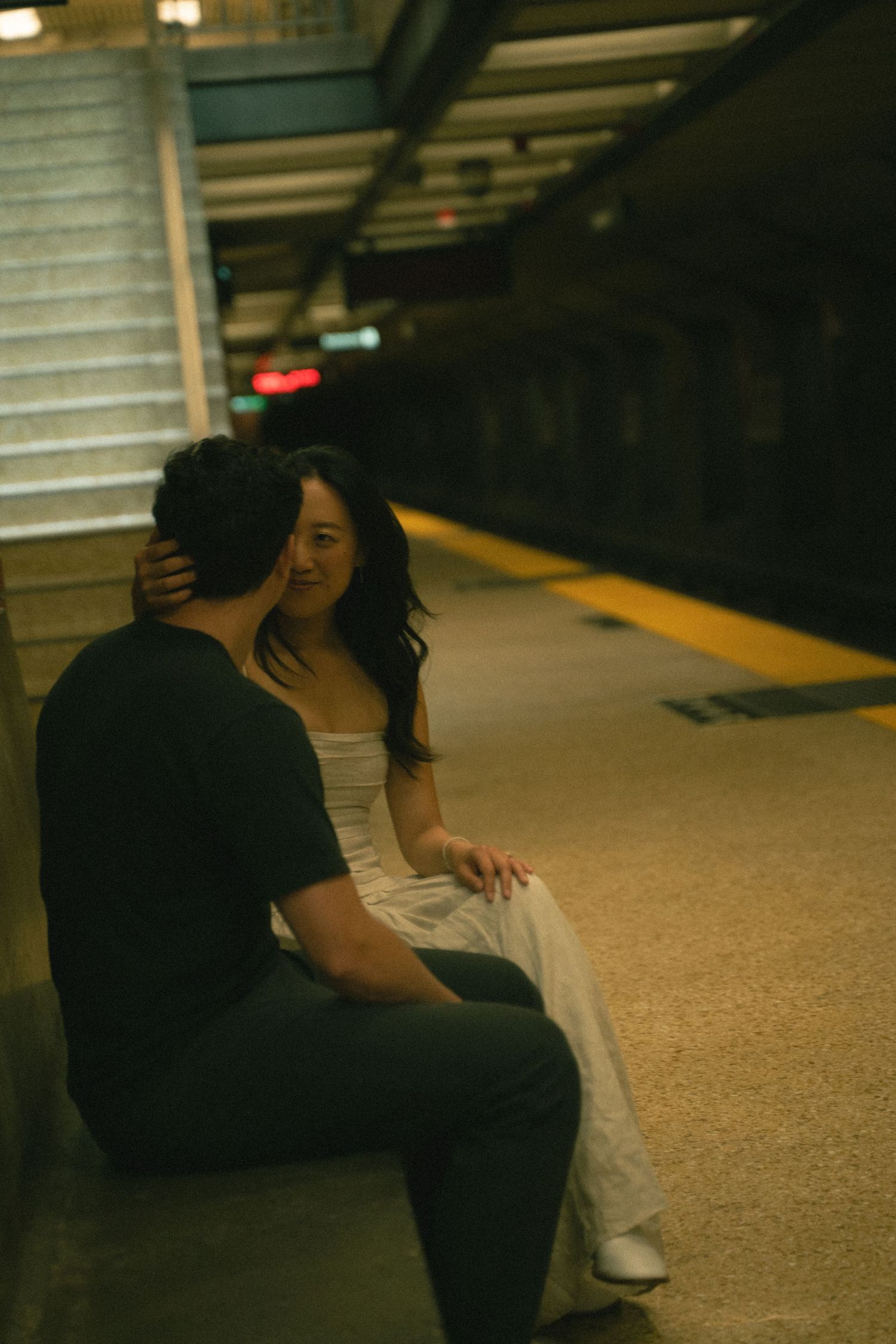 Couple sitting closely on a subway platform edge, sharing a quiet intimate moment together.