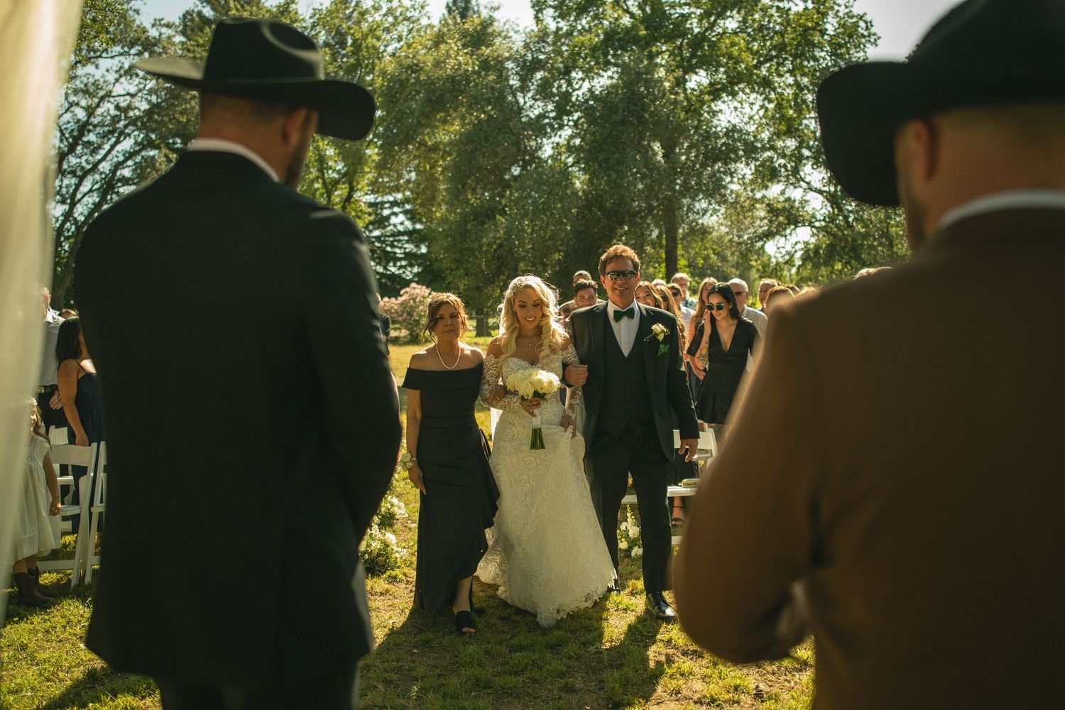 Bride walks down outdoor wedding aisle with escort amid guests in cowboy hats on sunny day.