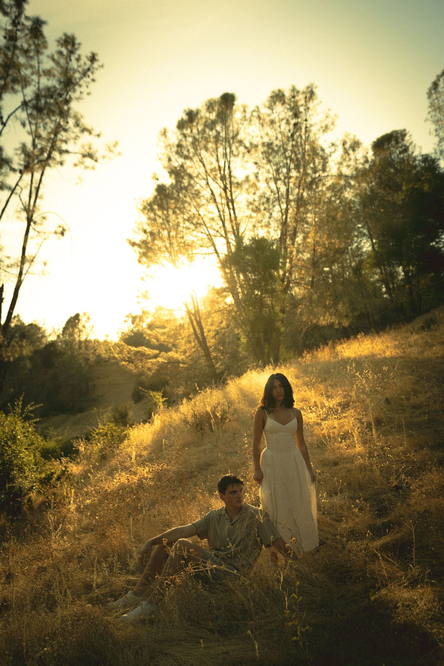 Couple in golden sunset light on a grassy hillside, woman in white dress standing, man sitting among dry grass with trees behind.
