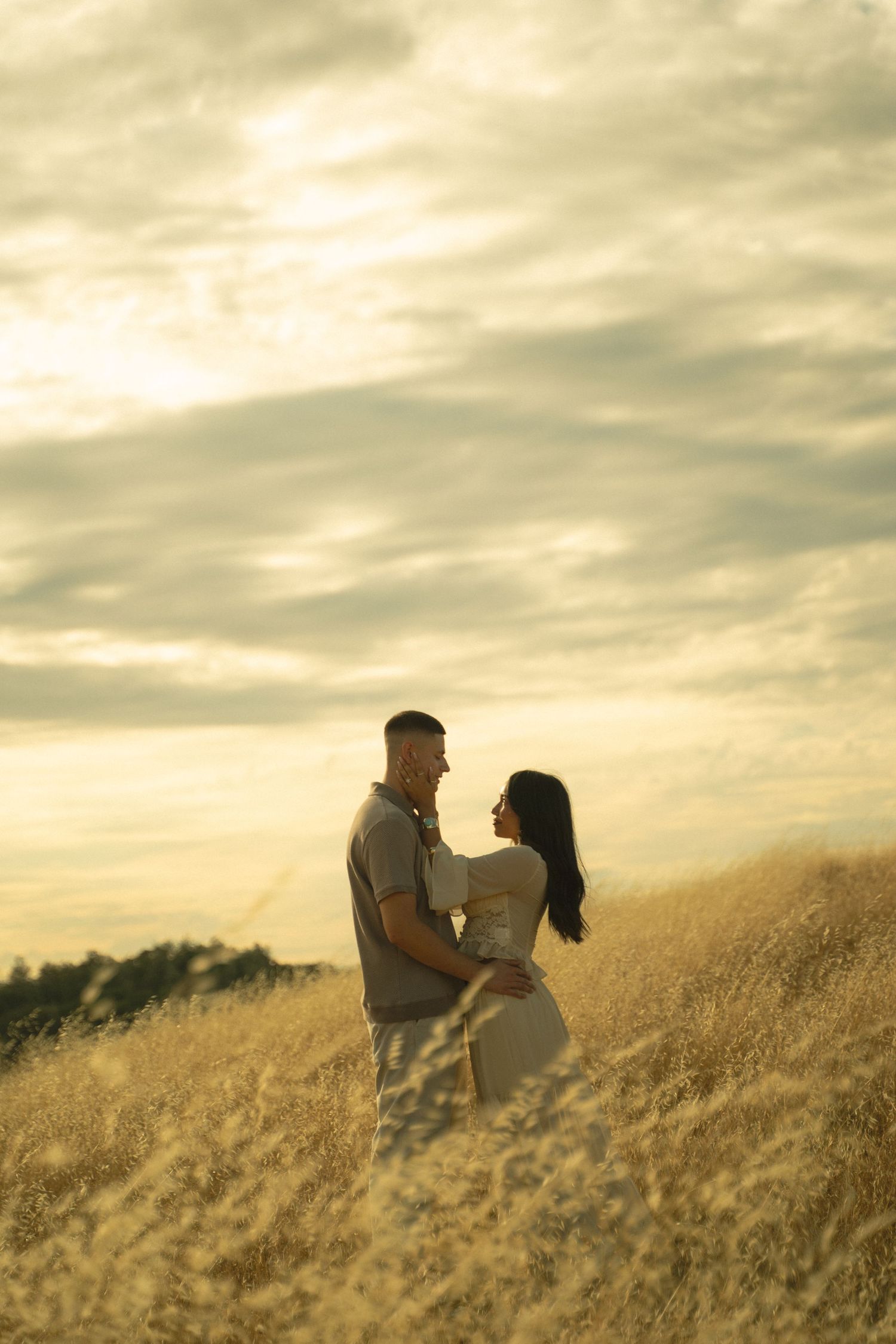 Couple embracing in golden field at sunset with dramatic cloudy sky.