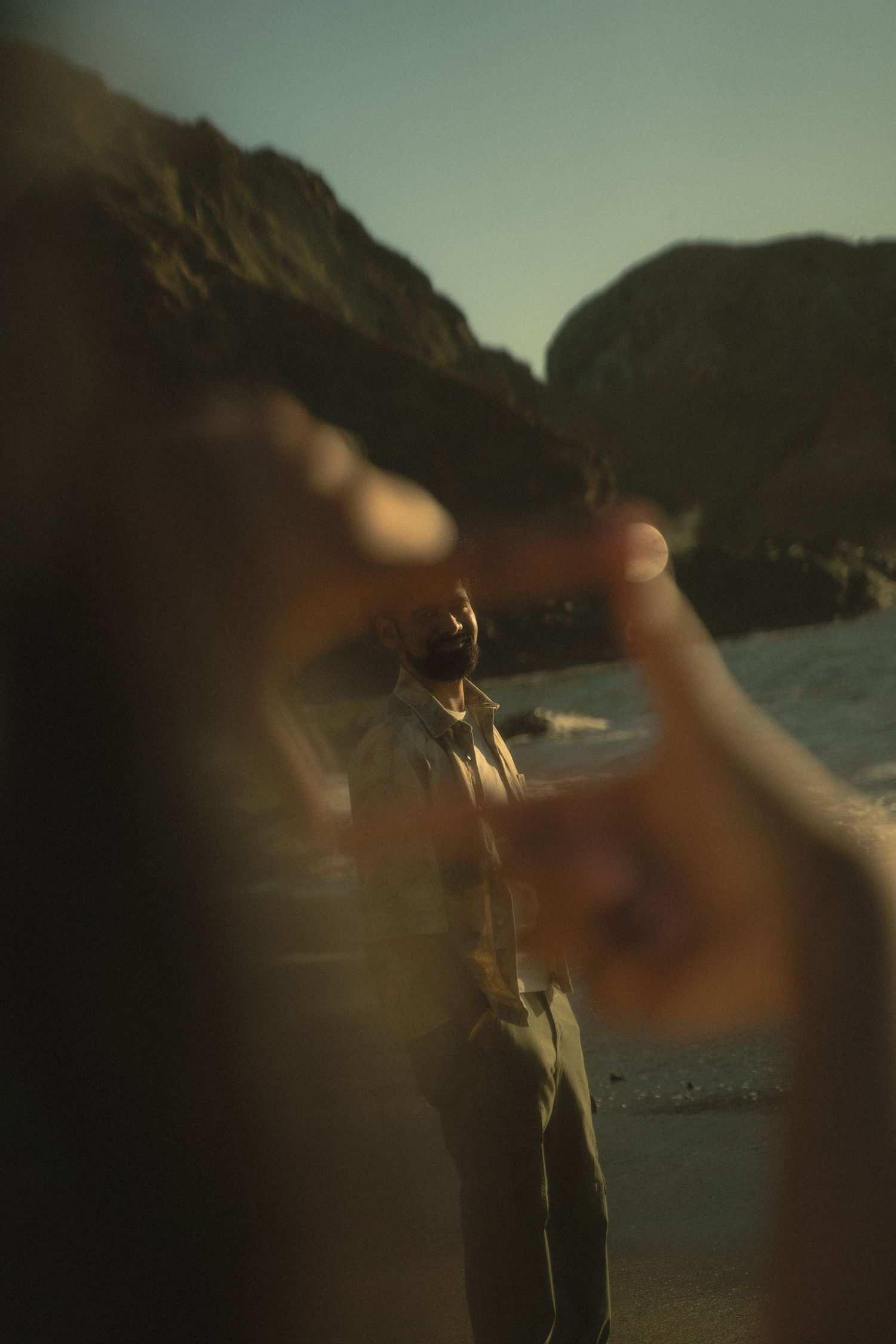 A person stands on a rocky beach framed by blurred hands forming a viewfinder gesture at golden hour.