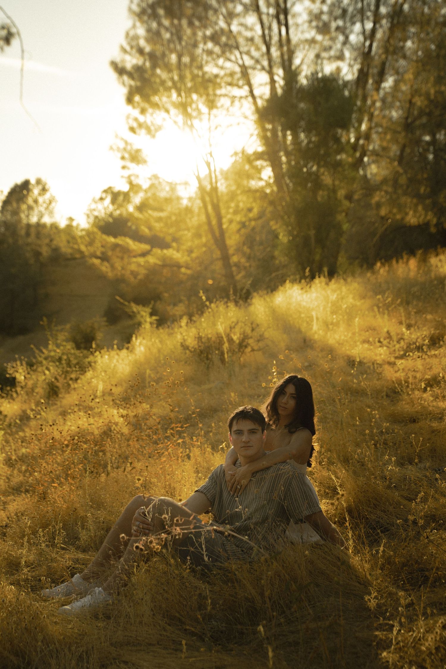 Couple sitting in golden sunlit meadow at sunset, surrounded by tall grass and trees in warm autumn light.