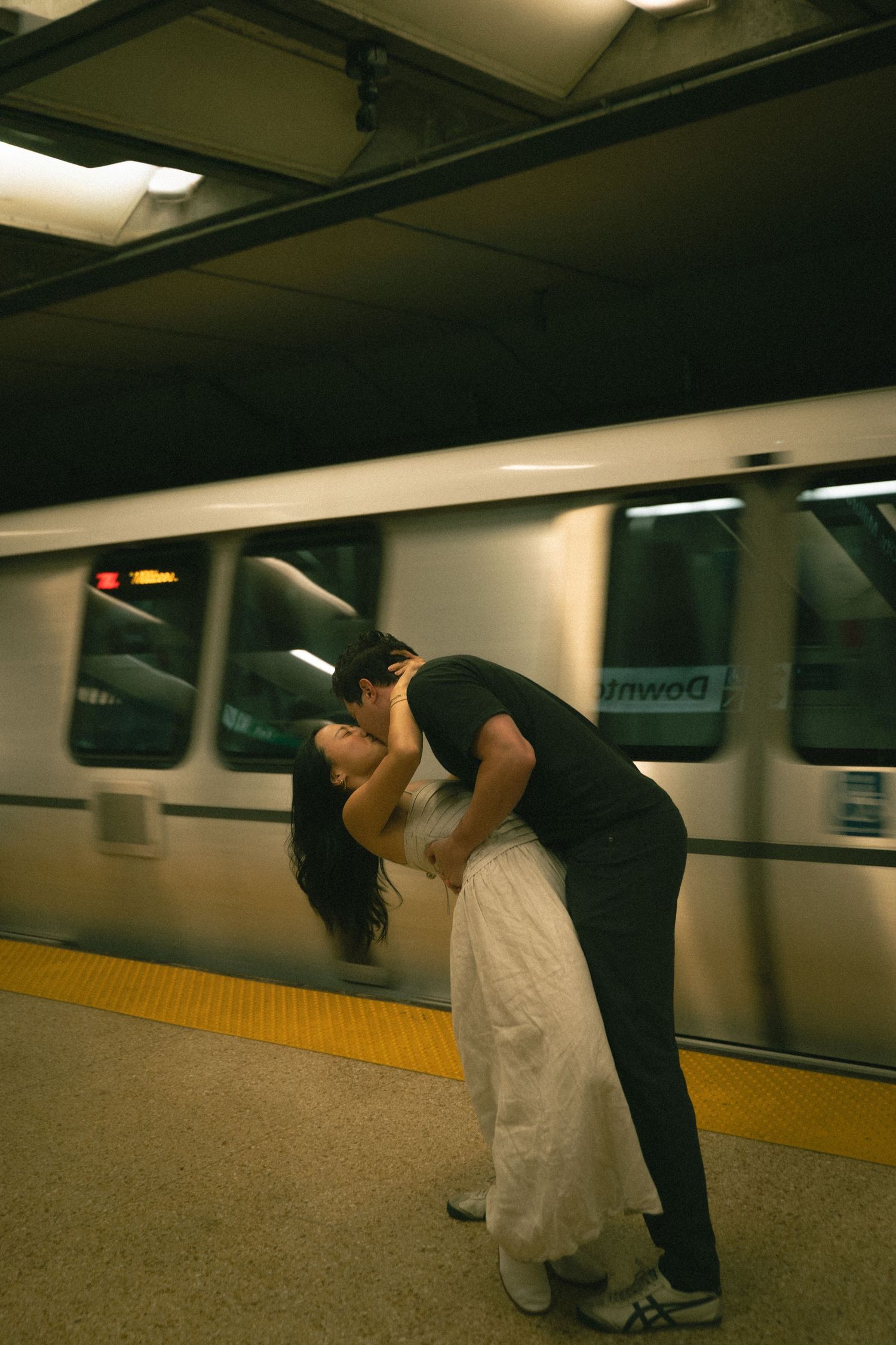 Couple sharing a passionate dip kiss on a subway platform with a moving train behind.