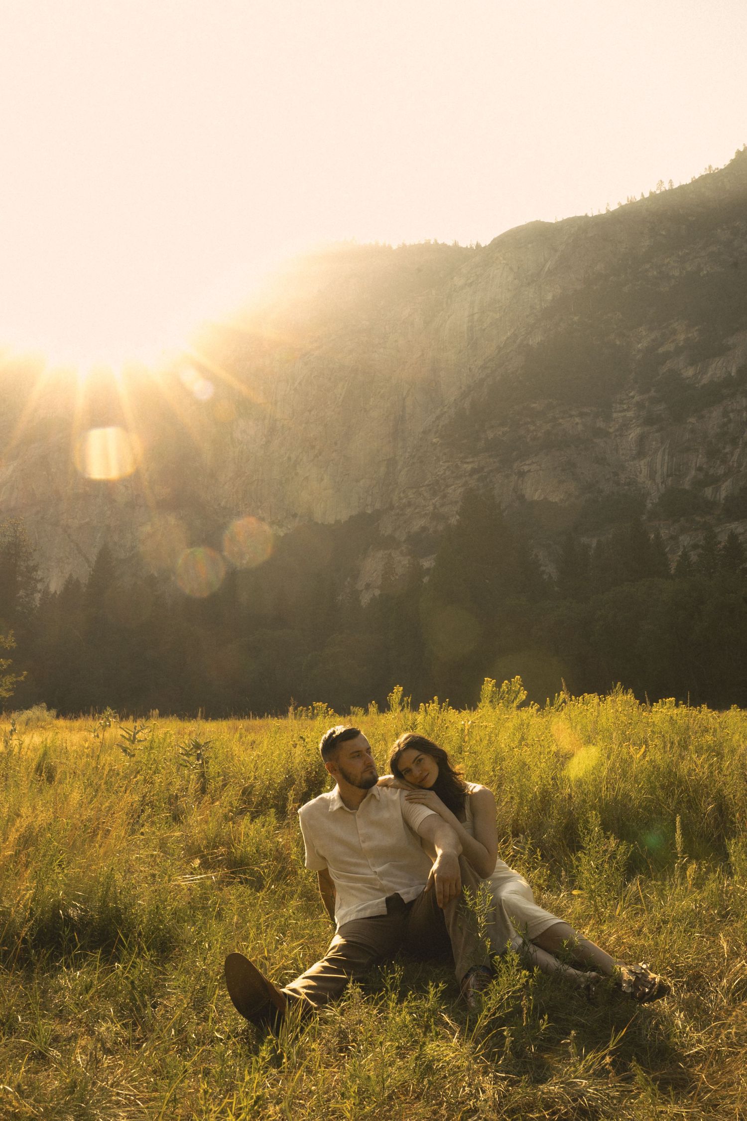 Couple sits together in sun-drenched meadow at golden hour with dramatic mountain cliffs behind them.
