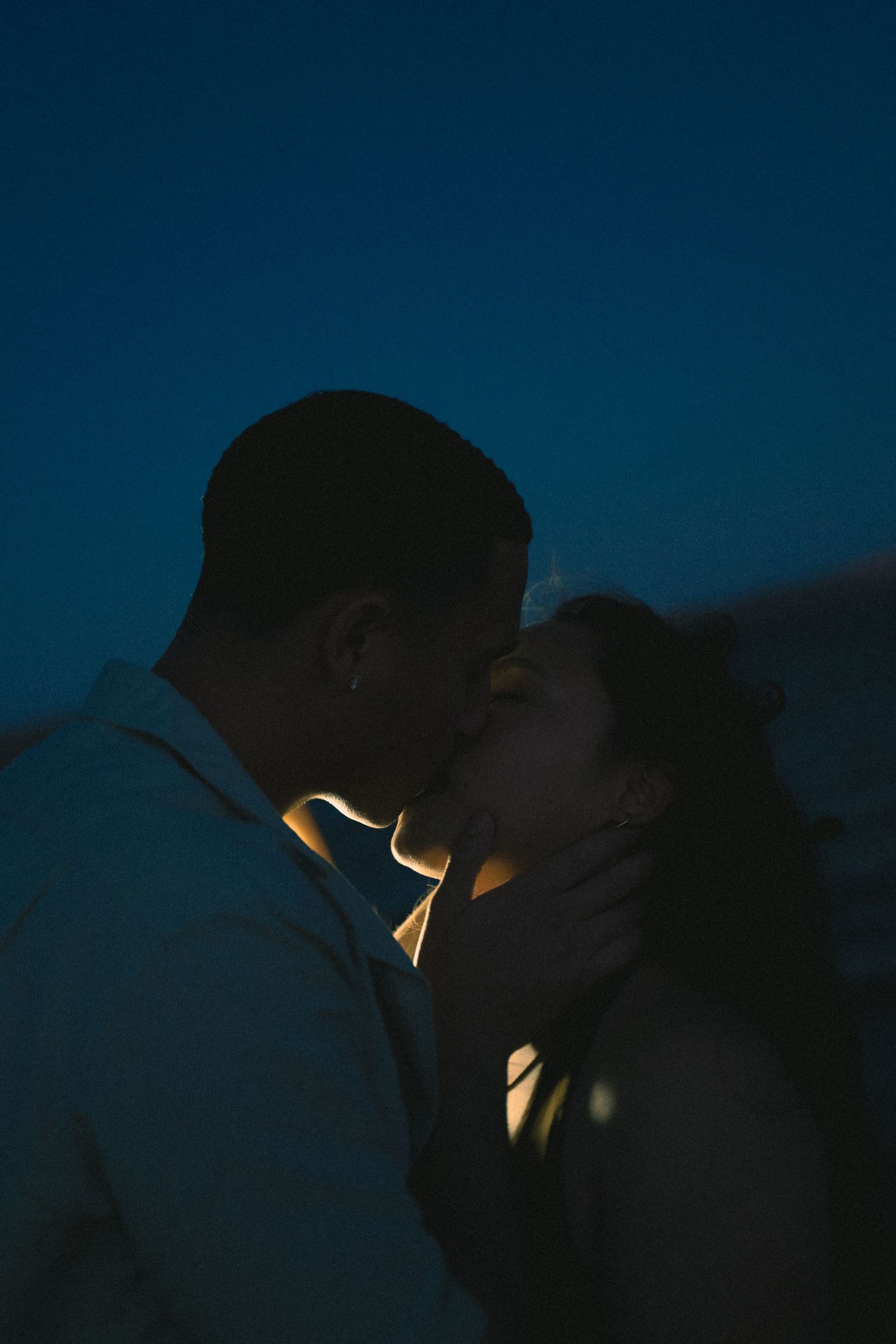 Close-up silhouette of couple sharing a kiss against a dark blue evening sky.