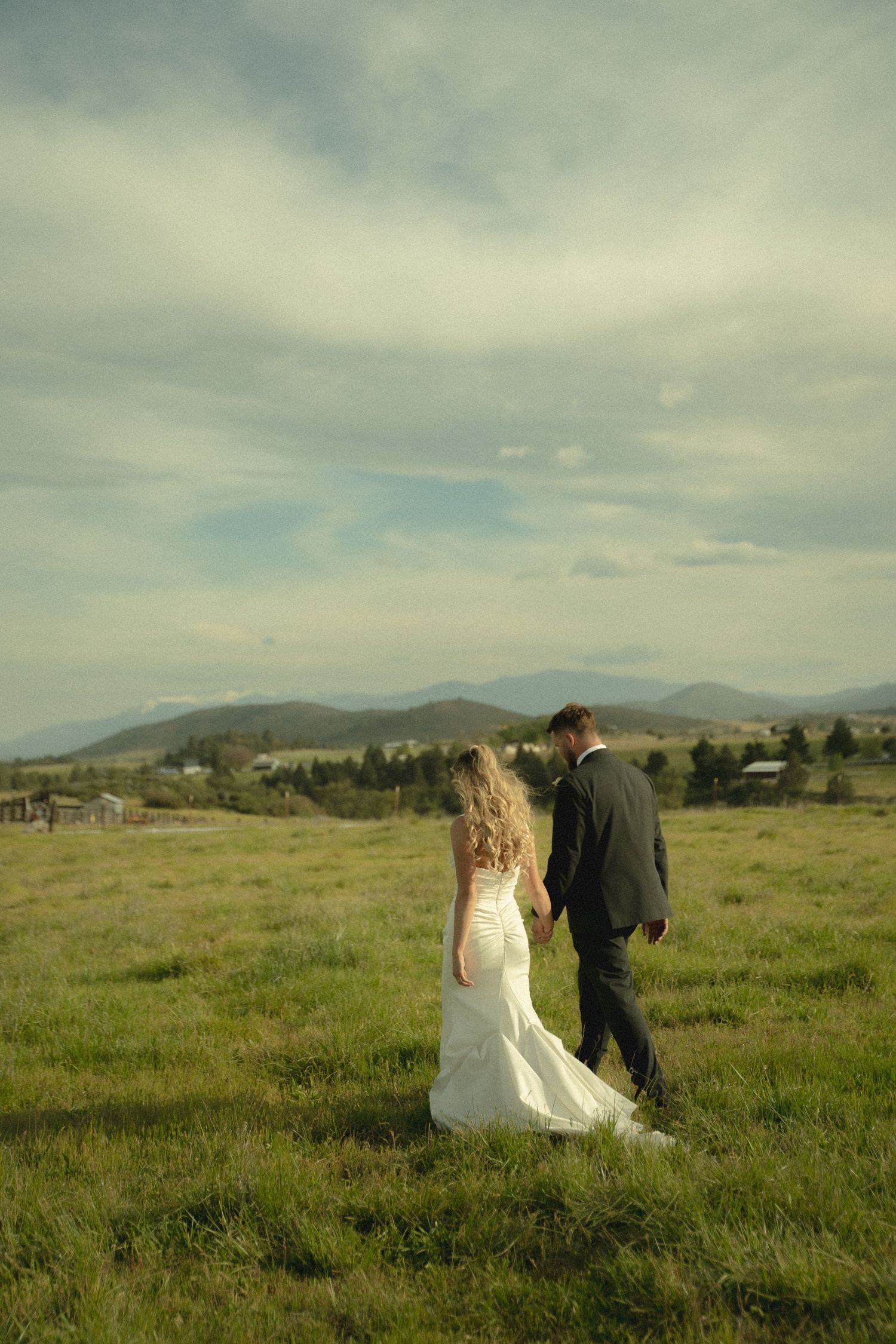Bride and groom walking hand-in-hand across a green meadow with mountain views.