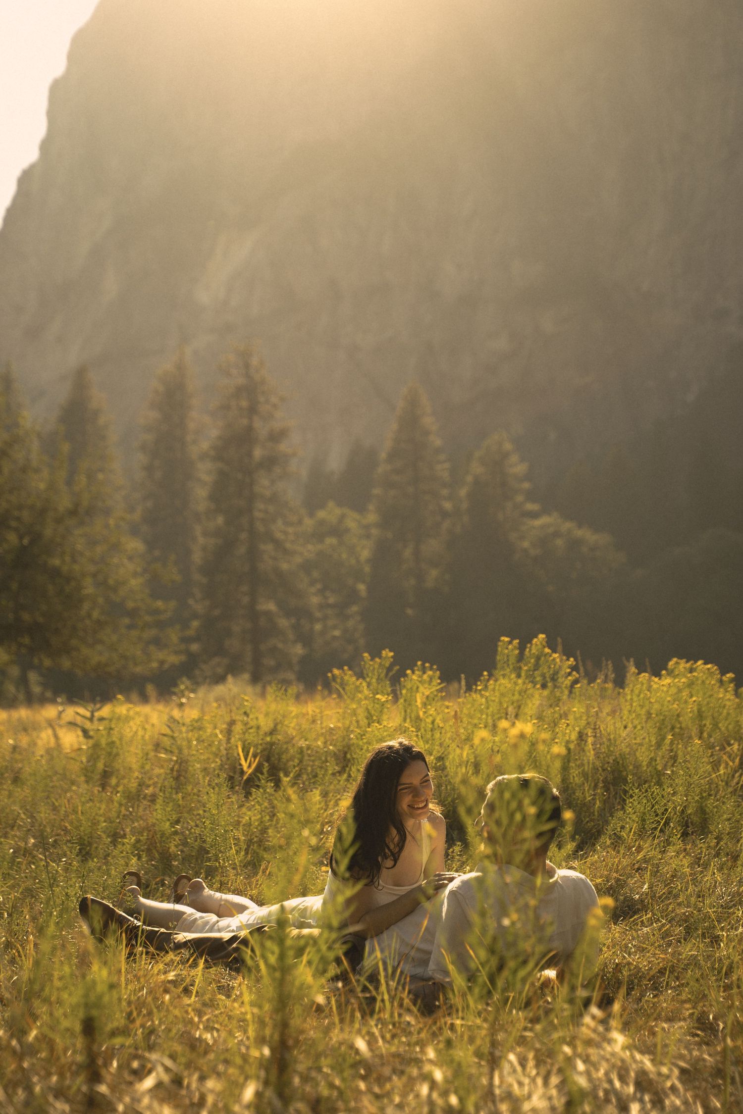 Couple sitting in golden sunlit meadow with tall pine trees and dramatic mountain backdrop during sunset.