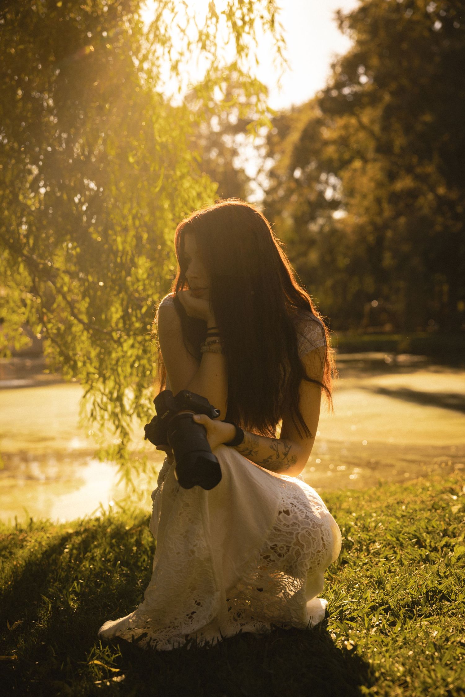 Woman in white lace dress kneeling by sunlit pond beneath weeping willow in golden hour light.