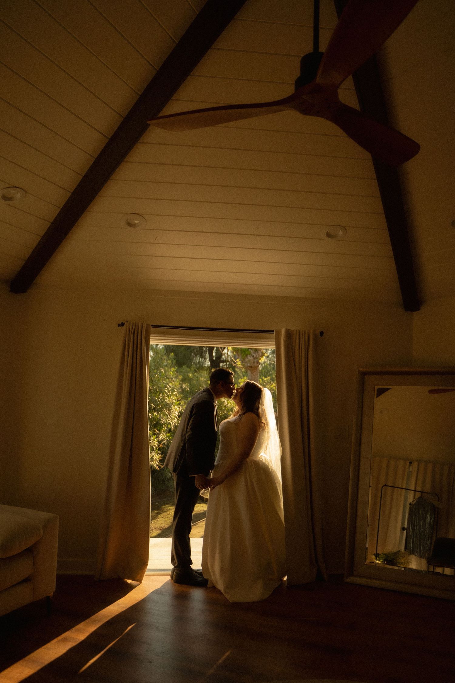 Bride and groom silhouetted in warm golden light kissing in a rustic room with vaulted ceiling.