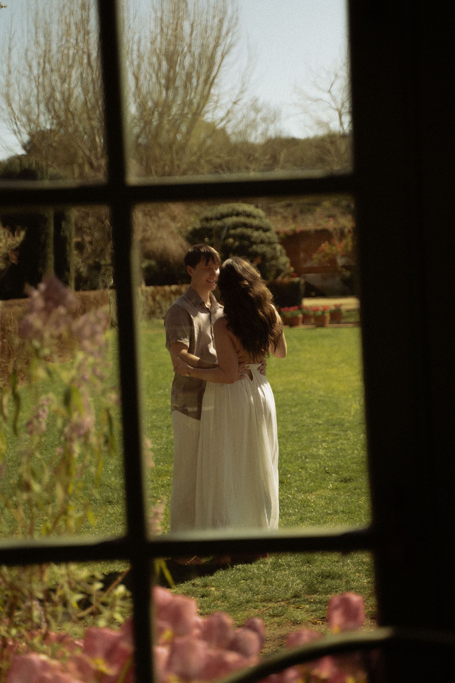 Couple embracing in a sunny garden viewed through a window frame with pink flowers.