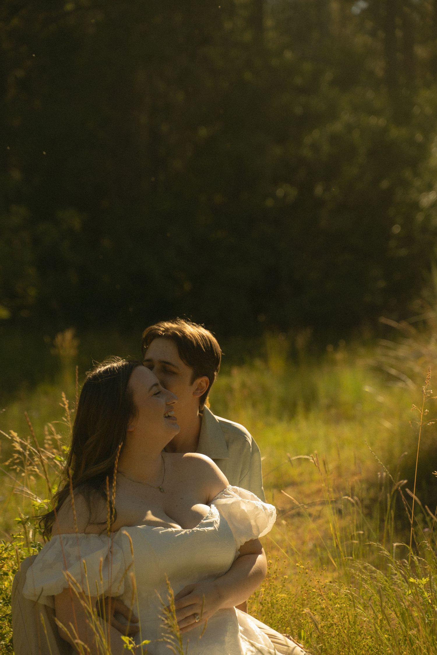 Couple embracing and kissing in golden sunlit grassy field surrounded by trees.