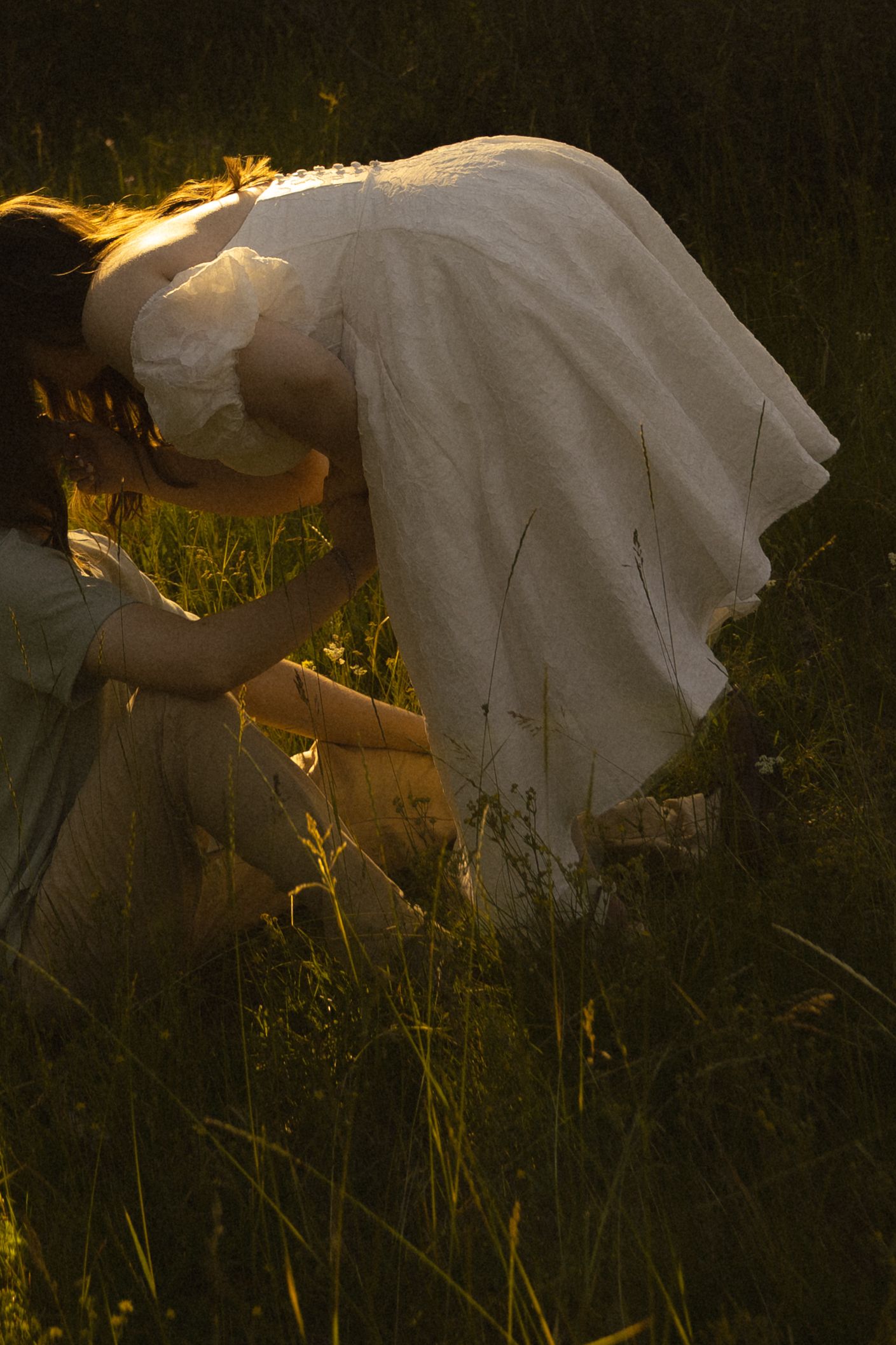 Woman in white dress kneeling in golden meadow grass at sunset.