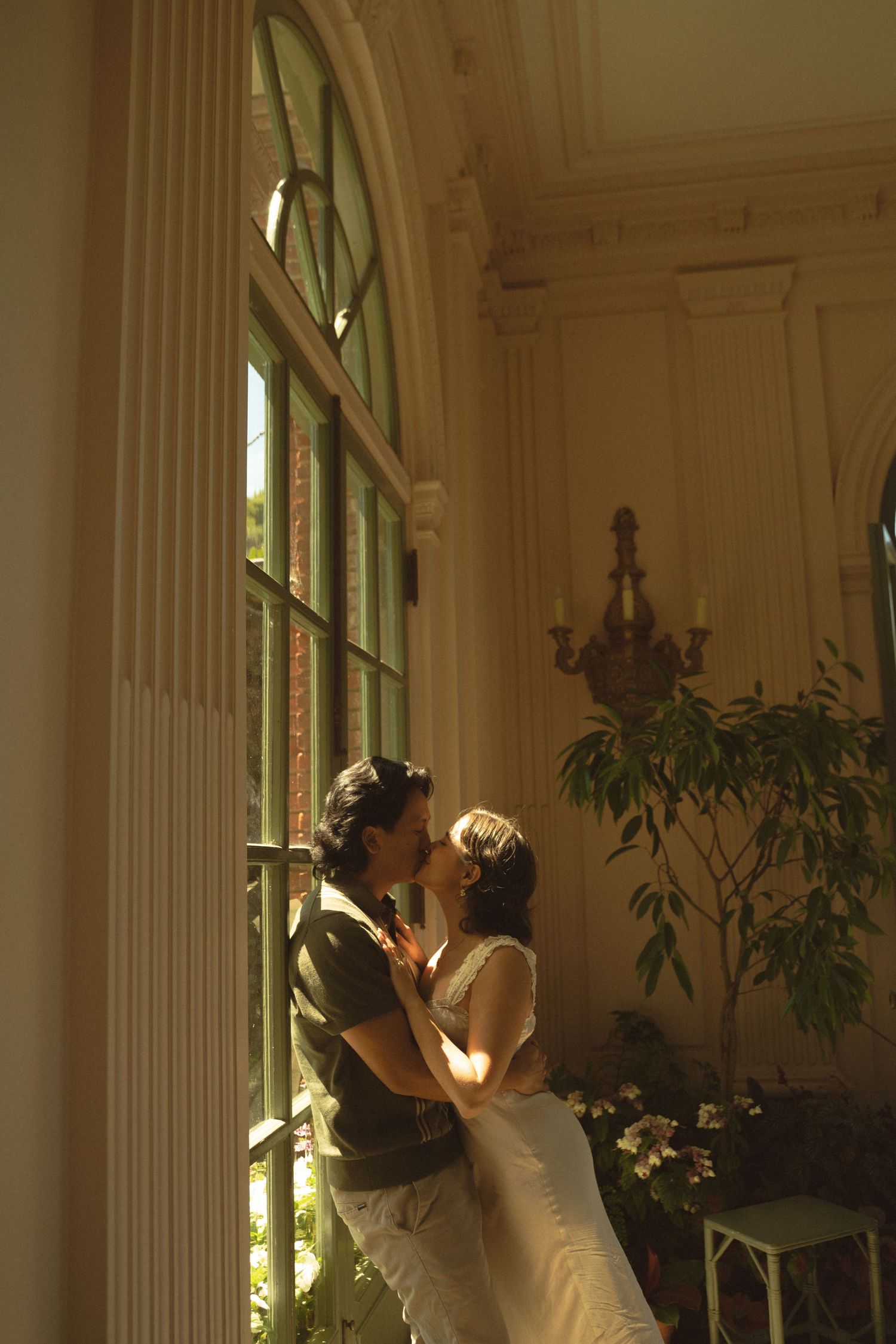 Couple sharing a romantic kiss by tall arched windows in an elegant sunlit room with ornate decor.