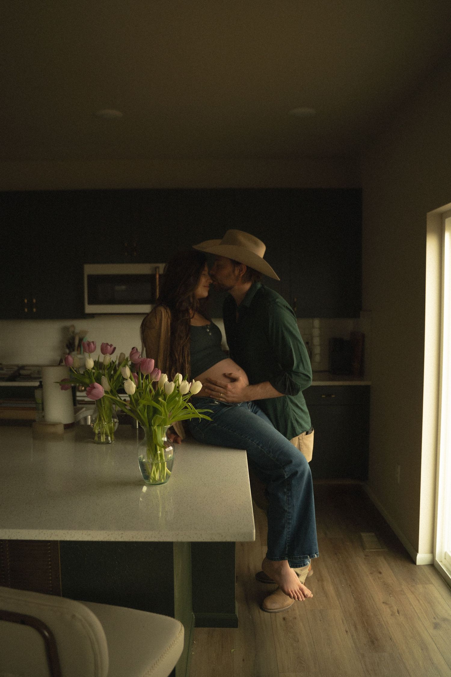 Couple embraces and kisses in a modern kitchen with tulips, one wearing a cowboy hat.