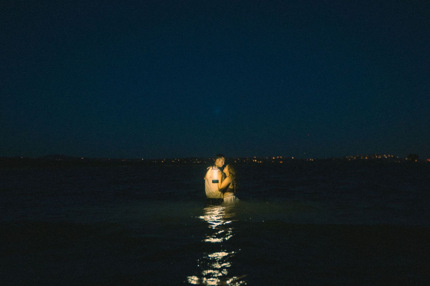 Couple standing in ocean water at night holding a glowing lantern with city lights in distance.