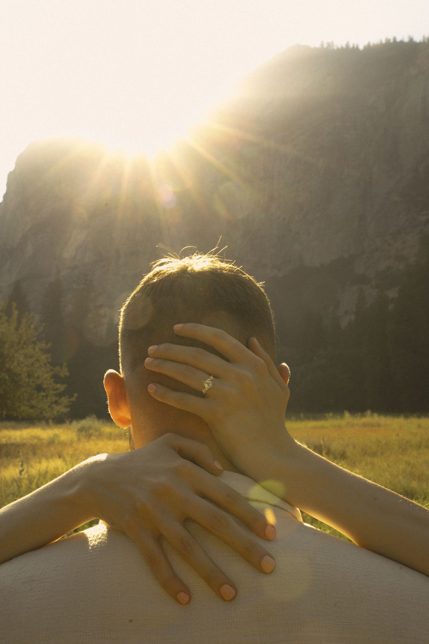 Close-up of a woman's hands with engagement ring embracing a man's neck against a sunny mountain meadow.