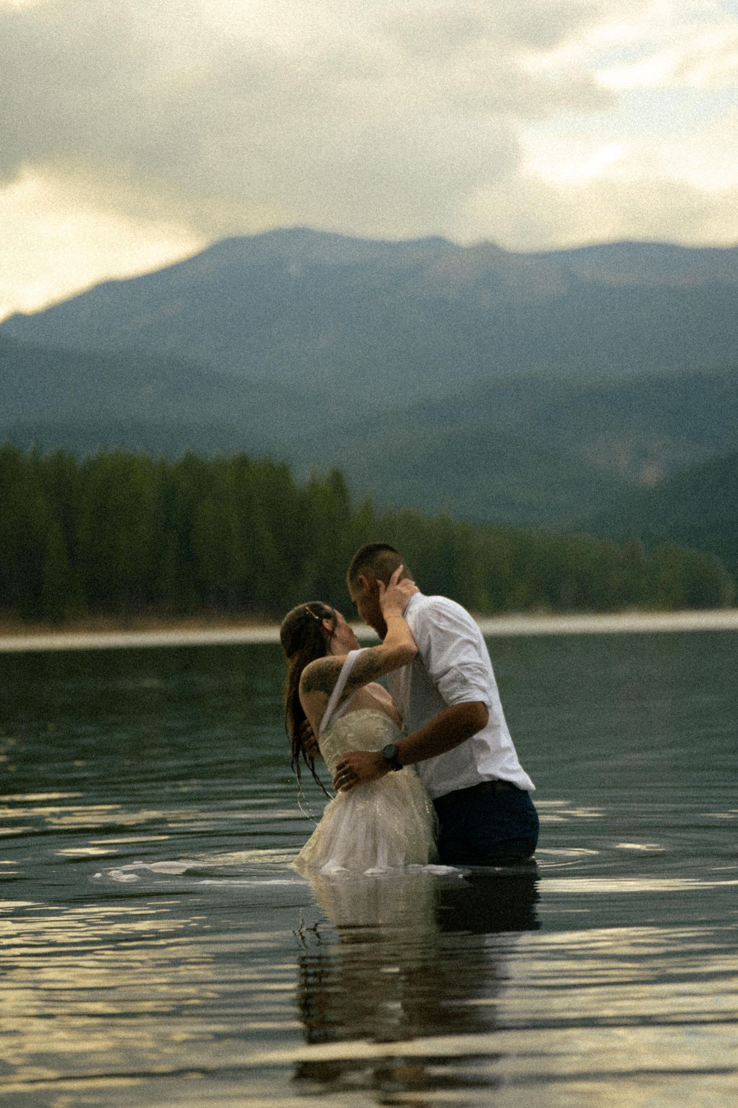 Couple kissing while standing in a mountain lake with forested hills and cloudy sky behind them.