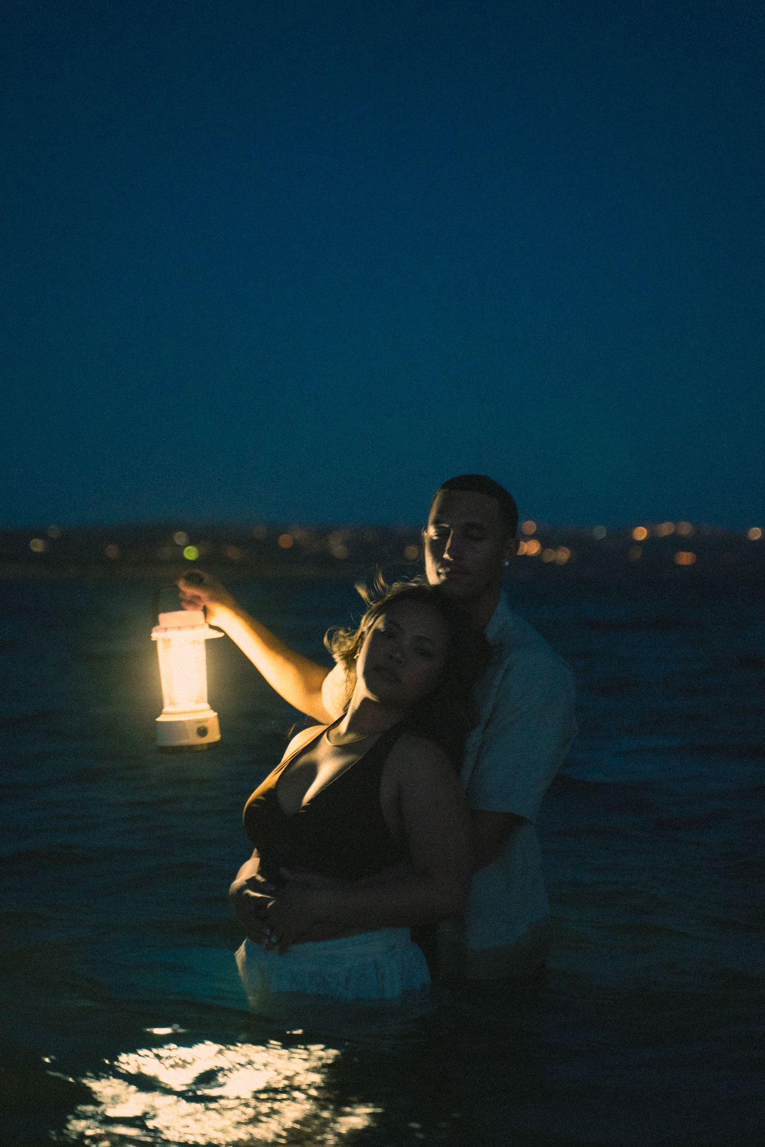Couple wading in ocean at night, woman holding a vintage glowing lantern with bokeh city lights.
