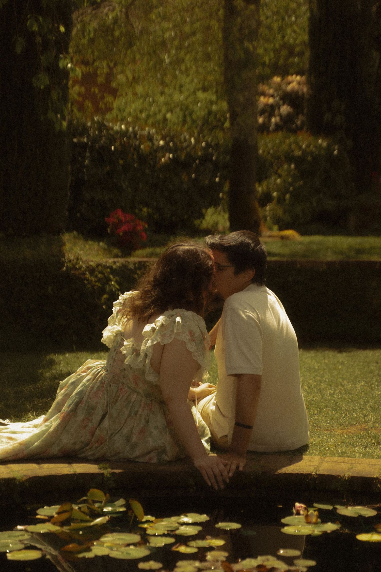 Couple shares a romantic kiss beside a lily pond in a sunlit garden with lush greenery.