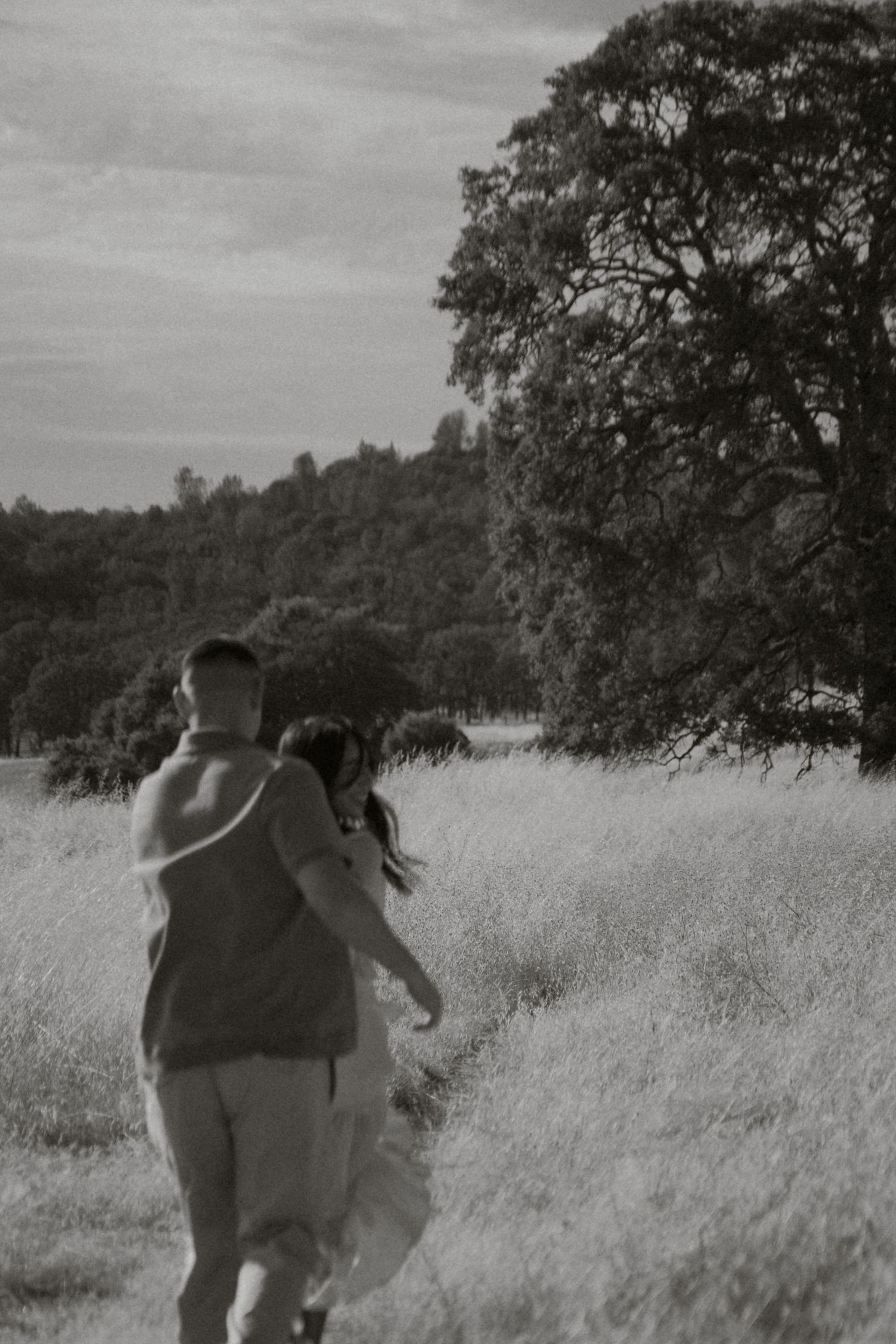 A couple walks hand in hand through a scenic meadow with tall trees in a black and white photo.