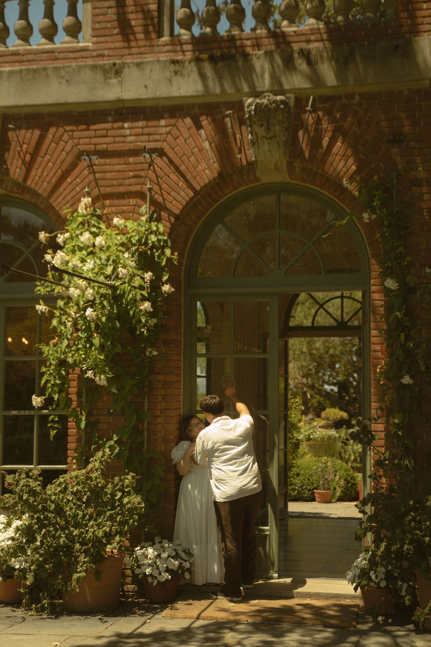 Couple embracing in arched doorway of ivy-covered brick mansion surrounded by lush garden greenery.