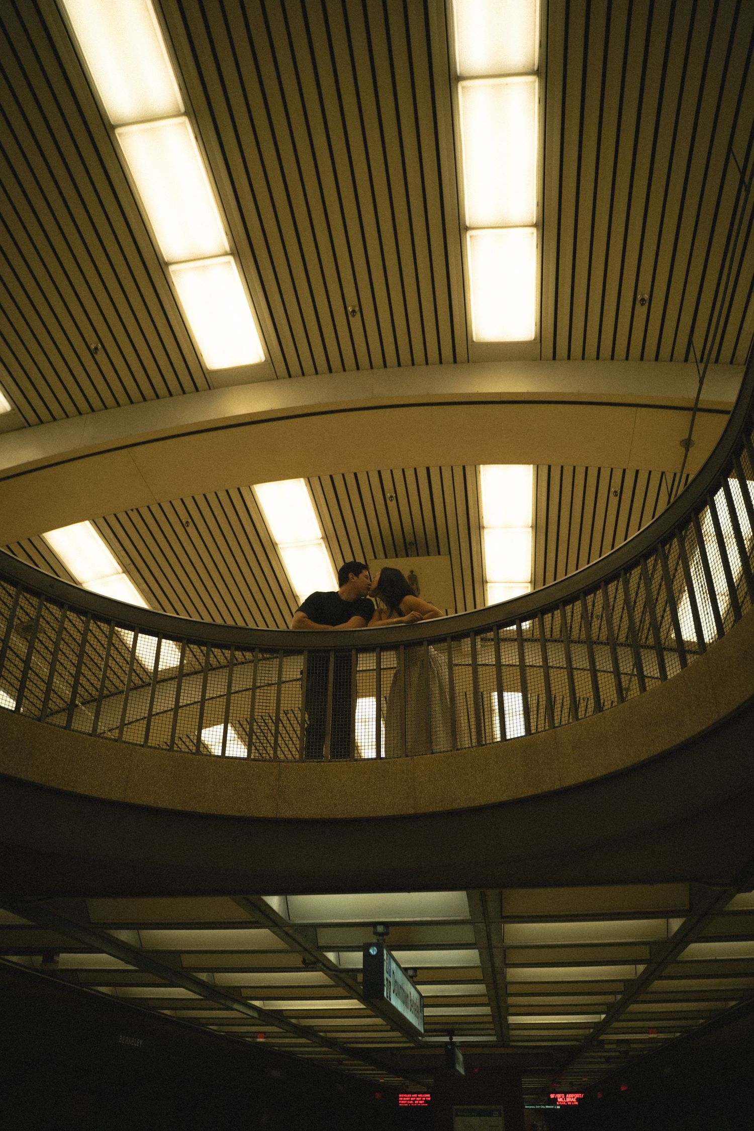 Couple kissing on a mezzanine level overlooking a transit station interior.