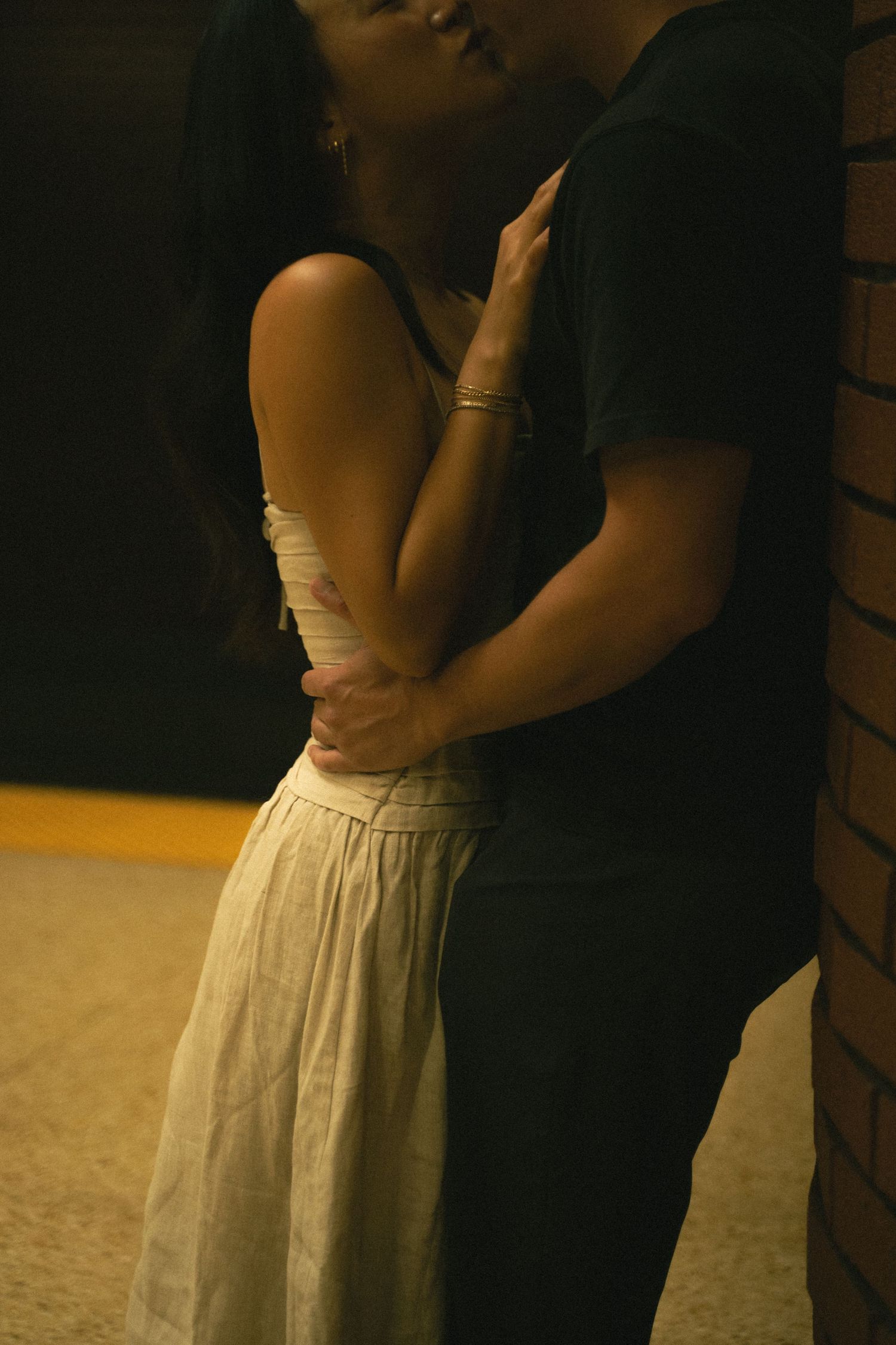 Couple sharing an intimate kiss against a brick wall in warm moody indoor lighting.