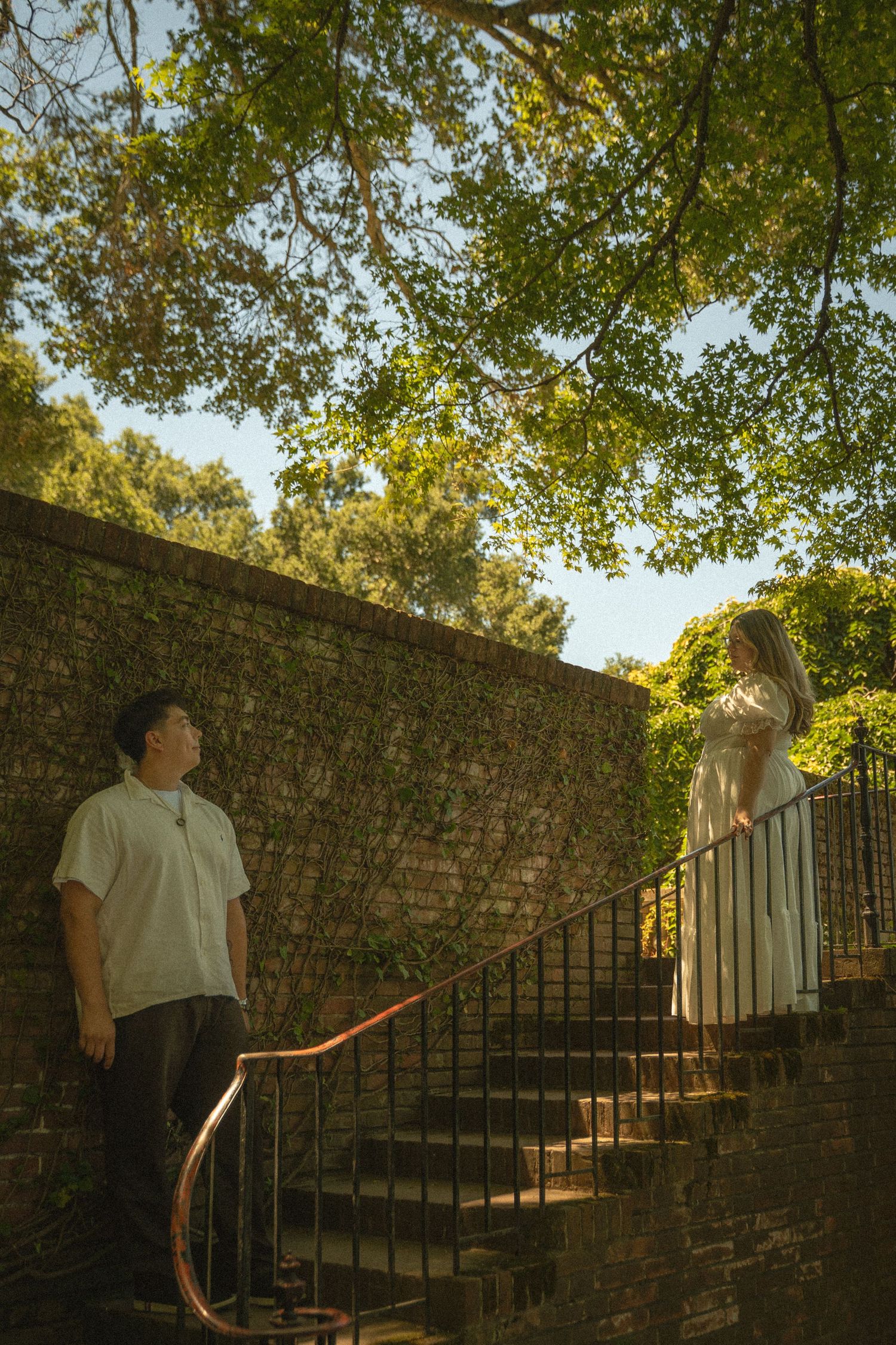 Couple on stone staircase outdoors, man looking up at woman in white dress beneath green trees.