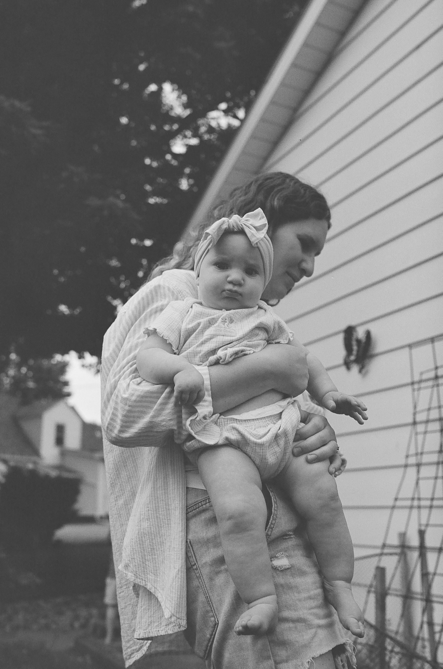 Mother holding baby girl in white bow headband outside home, black and white.