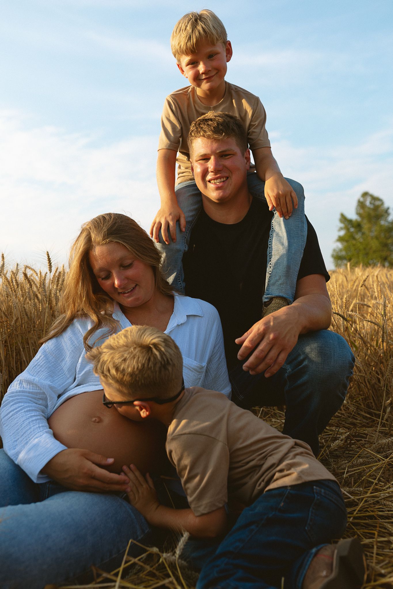 Family maternity photo with pregnant mom, dad, and two boys in a sunny wheat field.