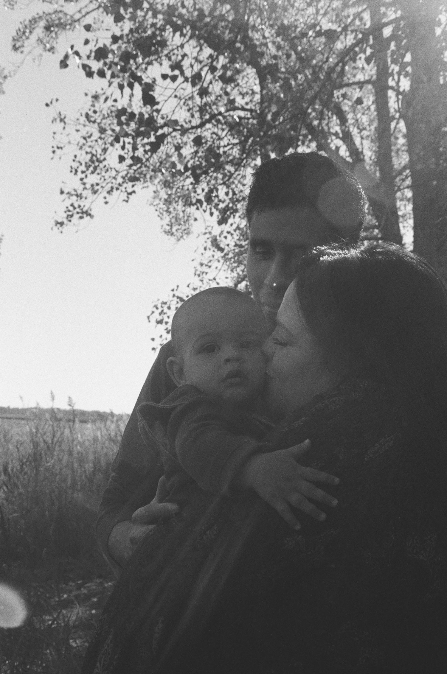Black and white photo of parents kissing their newborn baby outdoors near trees.
