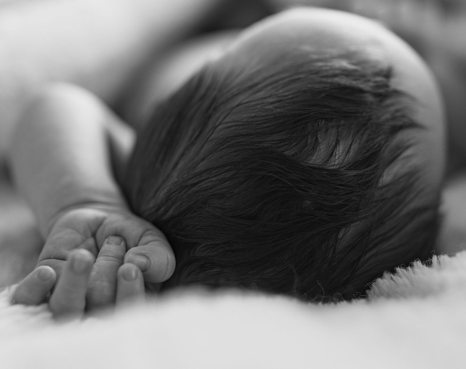 Black and white close-up of sleeping newborn baby with dark hair and tiny hand resting on soft blanket.