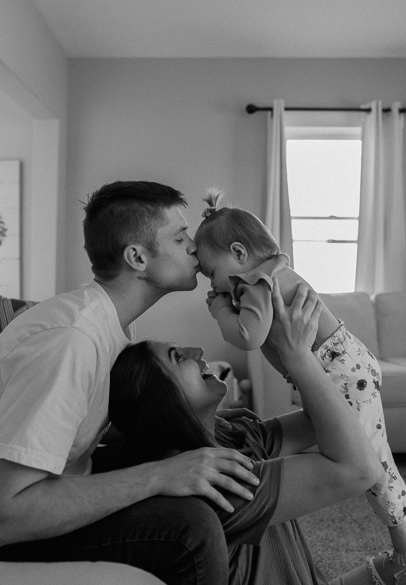 Black and white photo of young parents kissing their laughing baby together on living room couch at home.