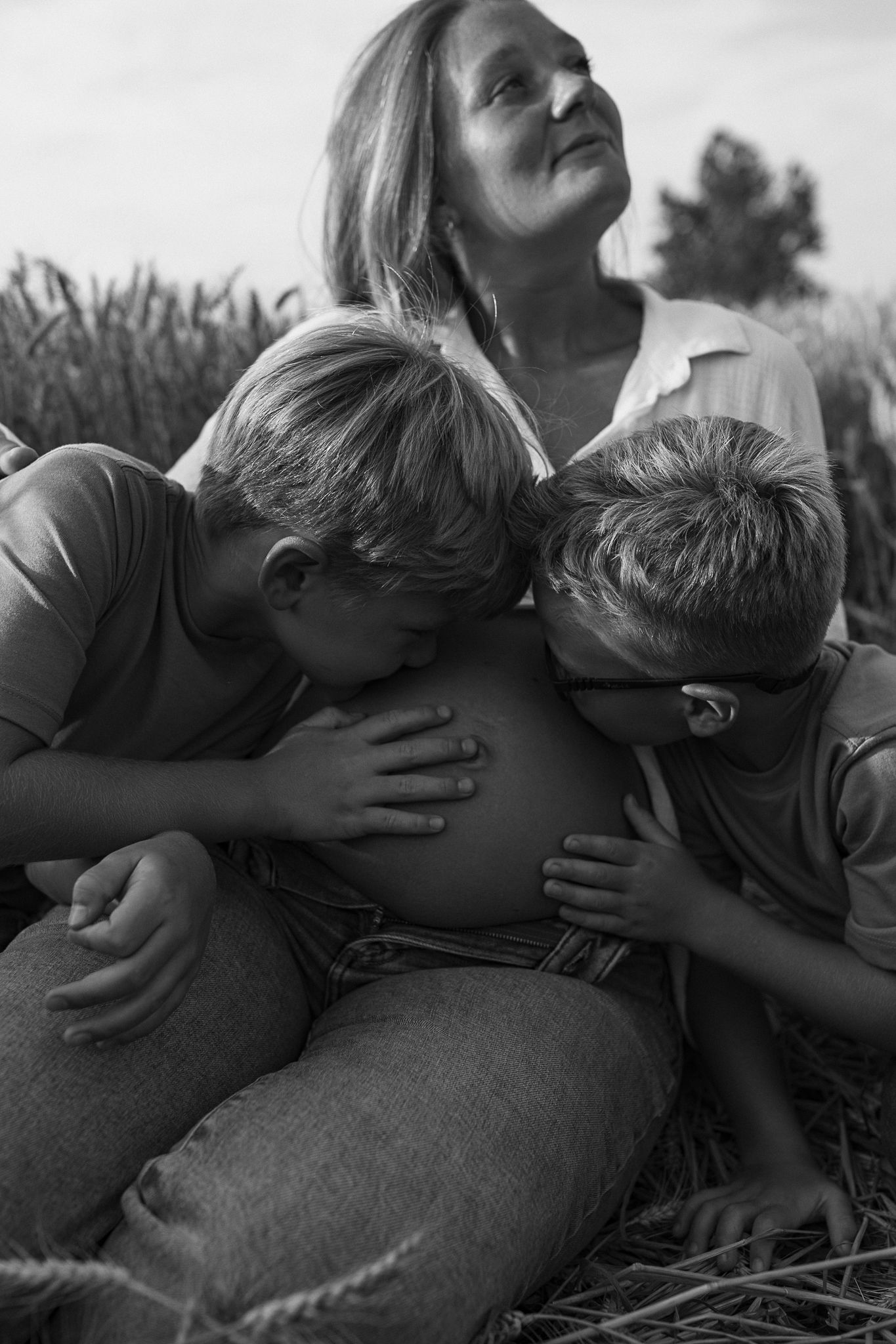Black and white image of two young boys kissing their pregnant mothers baby bump in a wheat field.