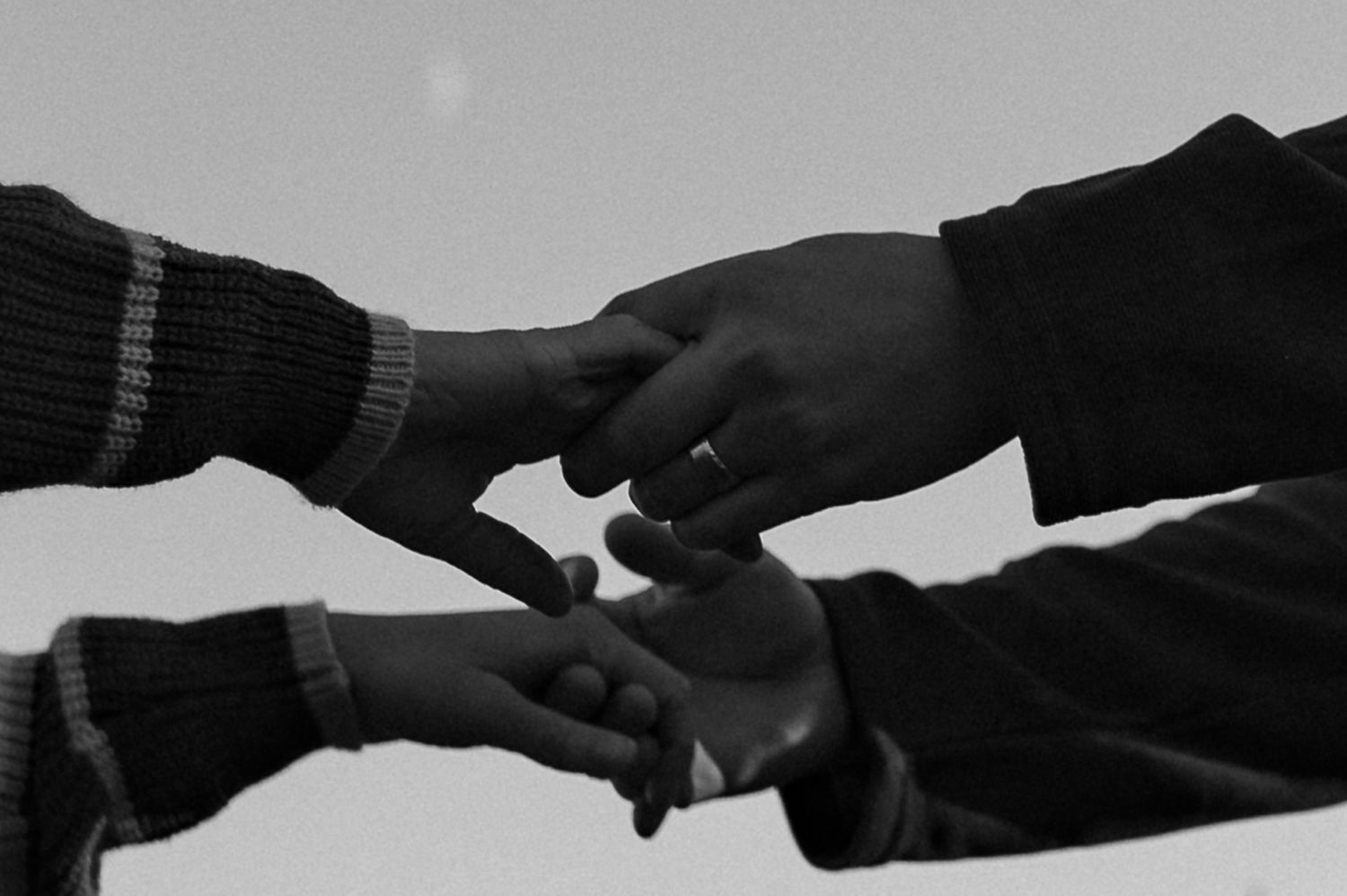 Black and white close-up of three people touching fingers together outdoors while wearing knit sweaters.