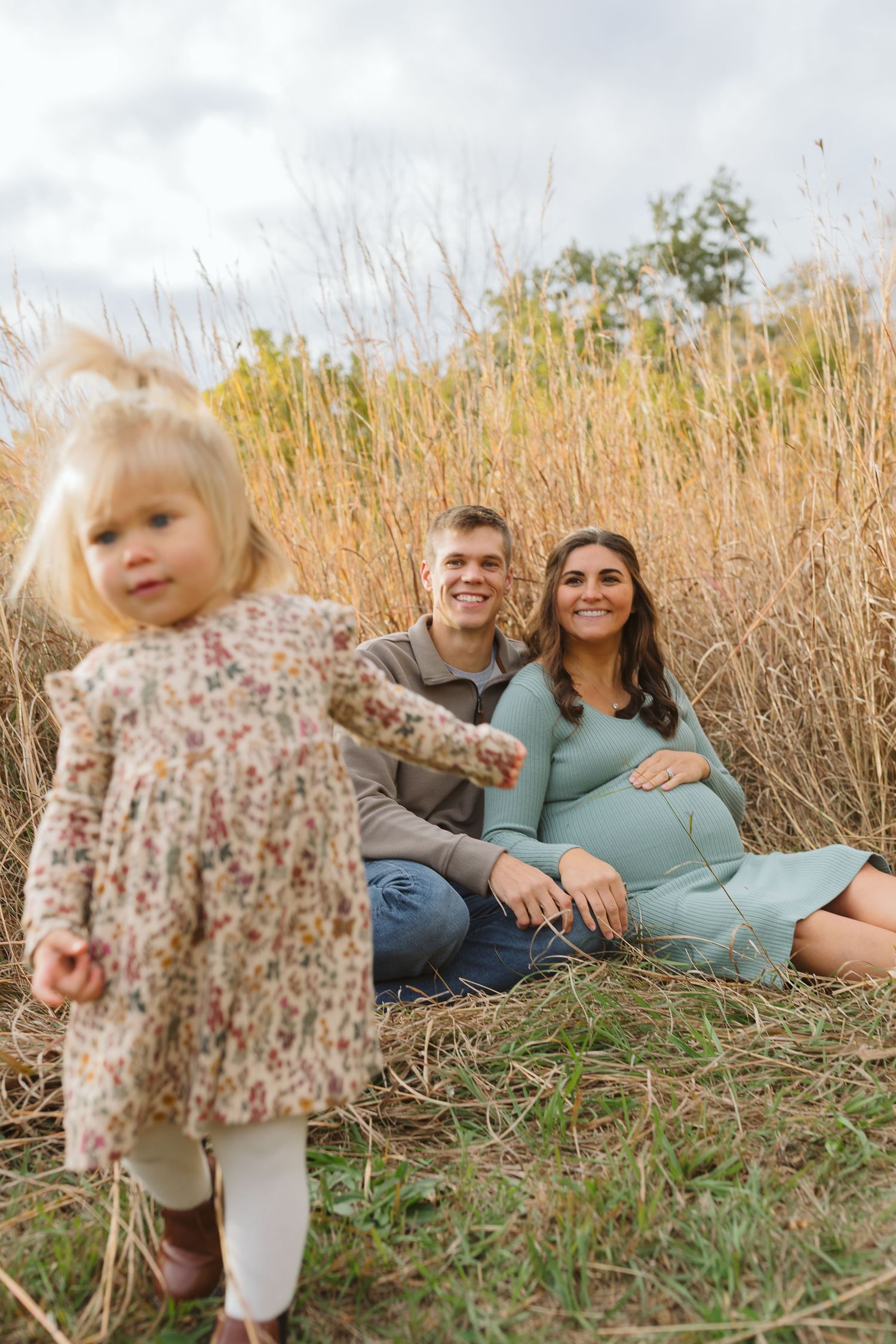 Smiling couple sitting in tall grass field with toddler girl in floral dress in foreground.