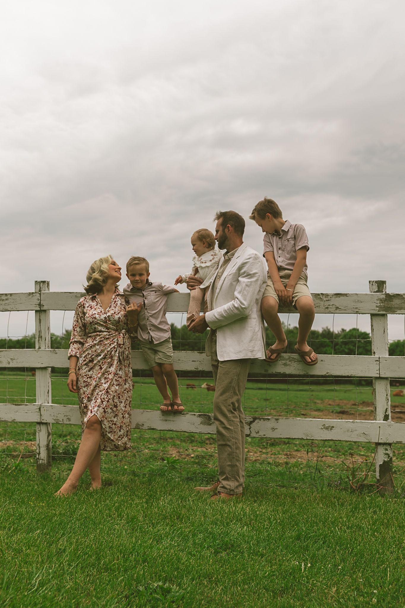 Family of five gathered around white wooden fence on green farm under overcast sky.