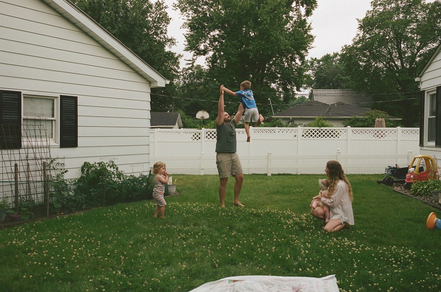 Father lifting child in backyard while mother holds baby near white fence.