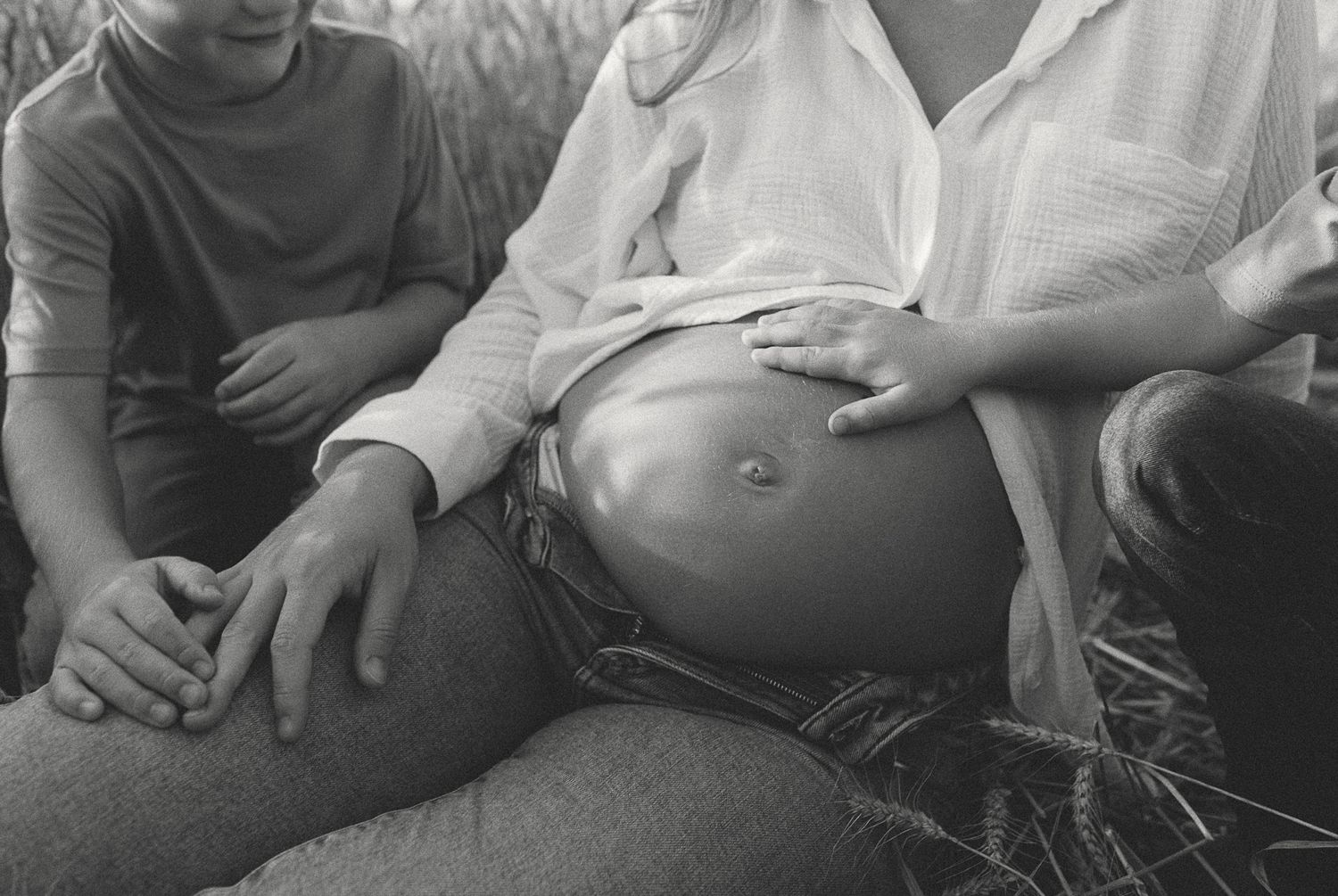 Black and white close-up of pregnant belly with child and partner hands resting on it outdoors.