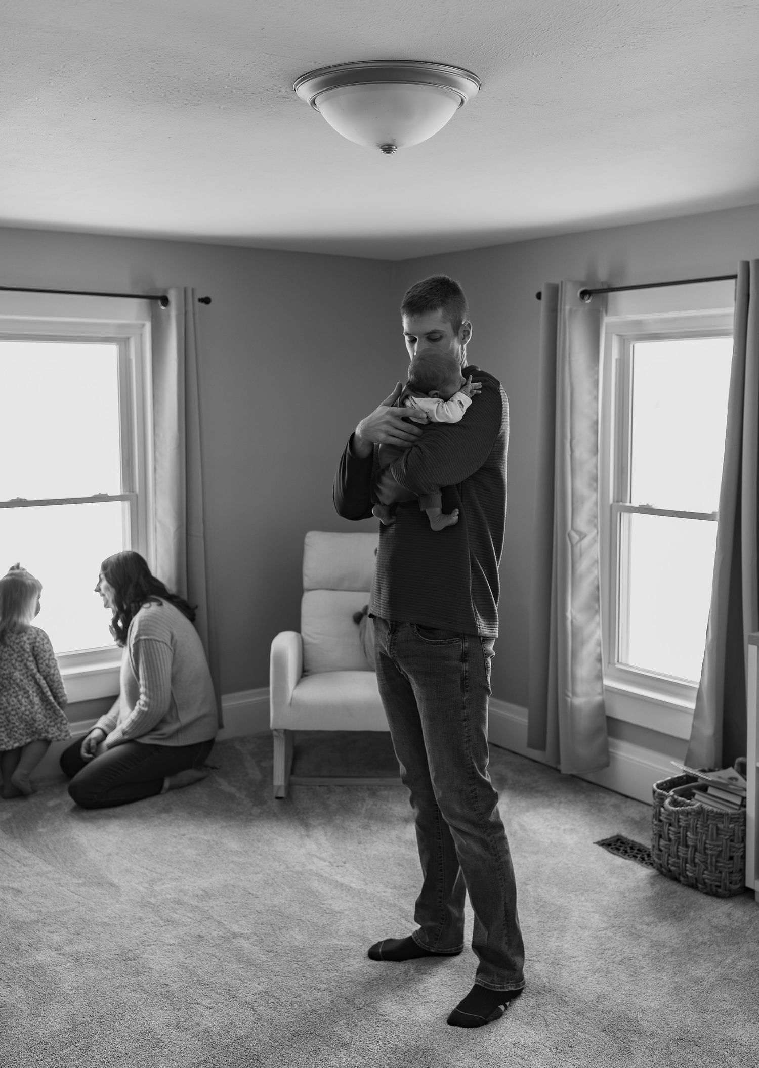 Black and white photo of father cradling newborn baby while mother and child sit by window in nursery.